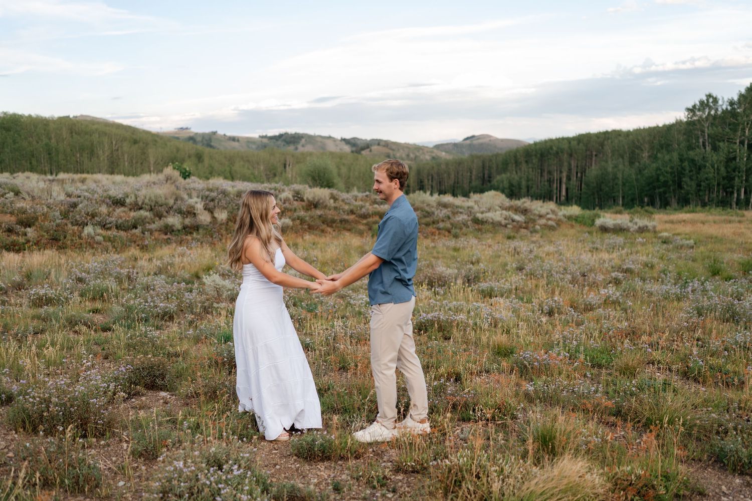 Couple holding hands in a scenic mountain meadow, woman in white dress, man in blue shirt, with forest backdrop.