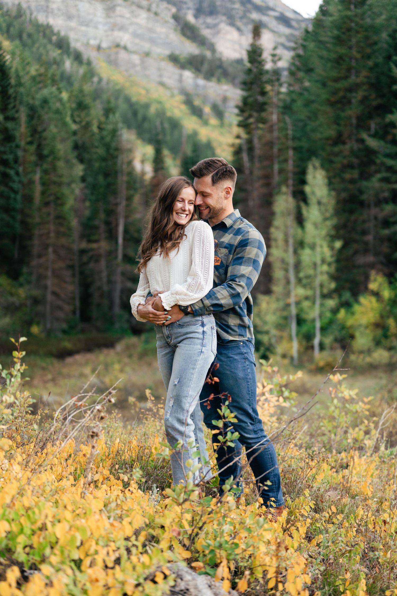 A couple embraces in a mountain meadow with evergreen trees and autumn colors surrounding them.