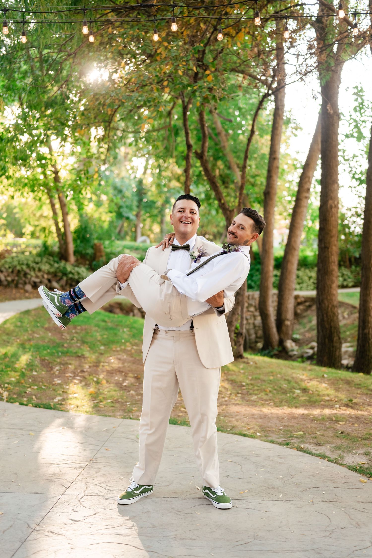 Two people in wedding attire share a joyful moment with one carrying the other on a garden path.