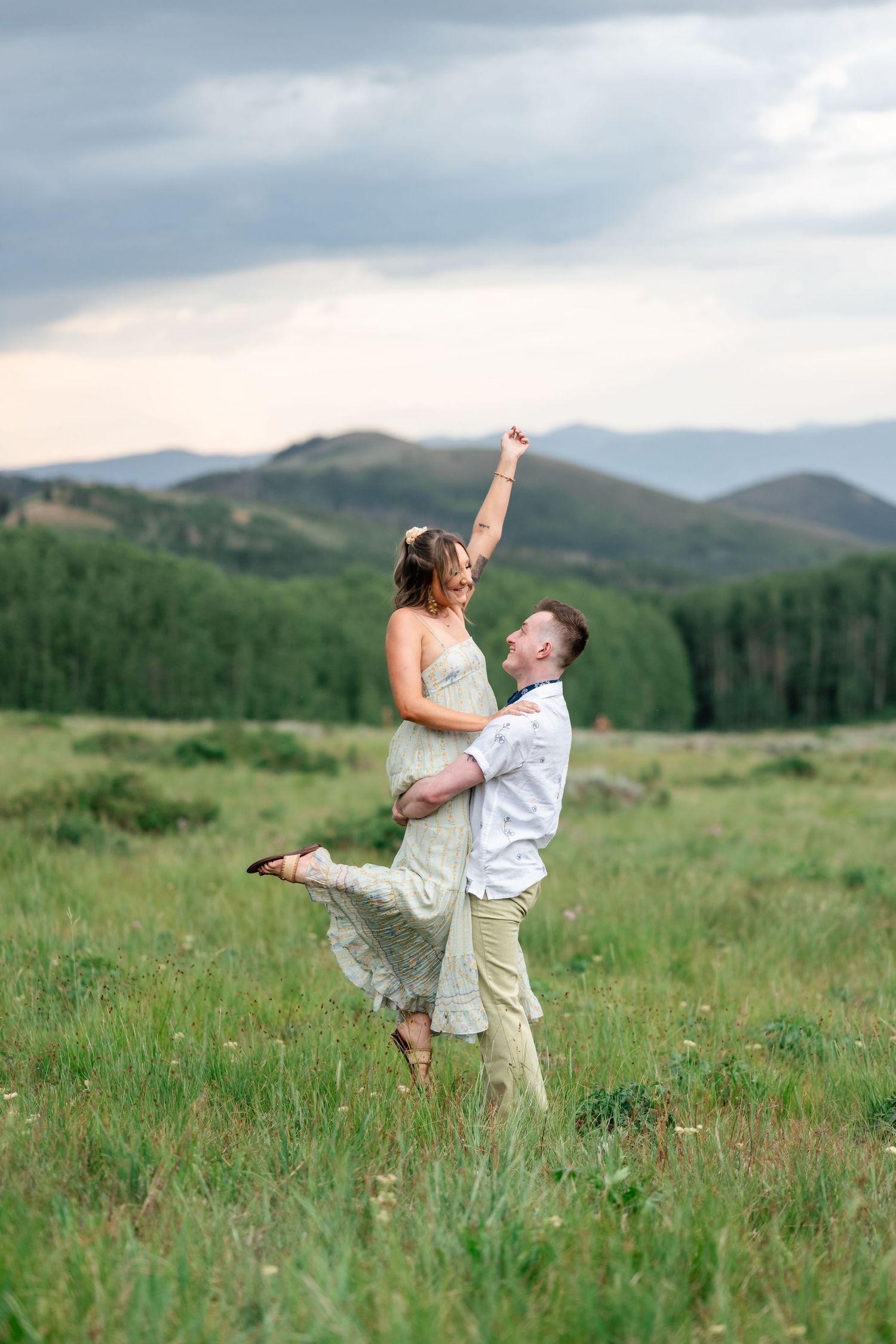 A romantic lift pose in a mountain meadow with dramatic cloudy skies and rolling hills in the background. Park City engagement photographer.