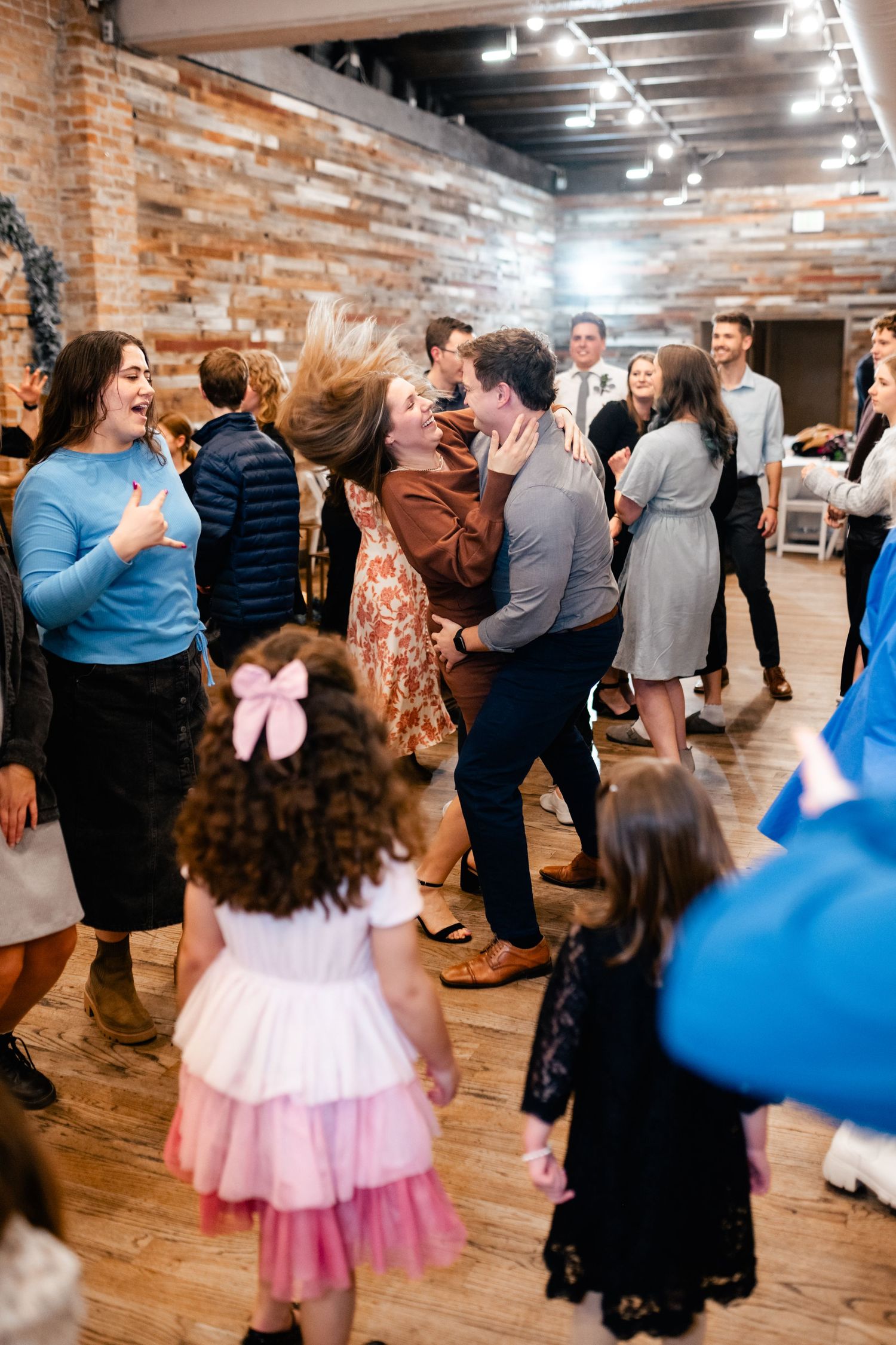 People dancing and celebrating at a party in a rustic brick-walled venue with wooden floors and overhead lighting.