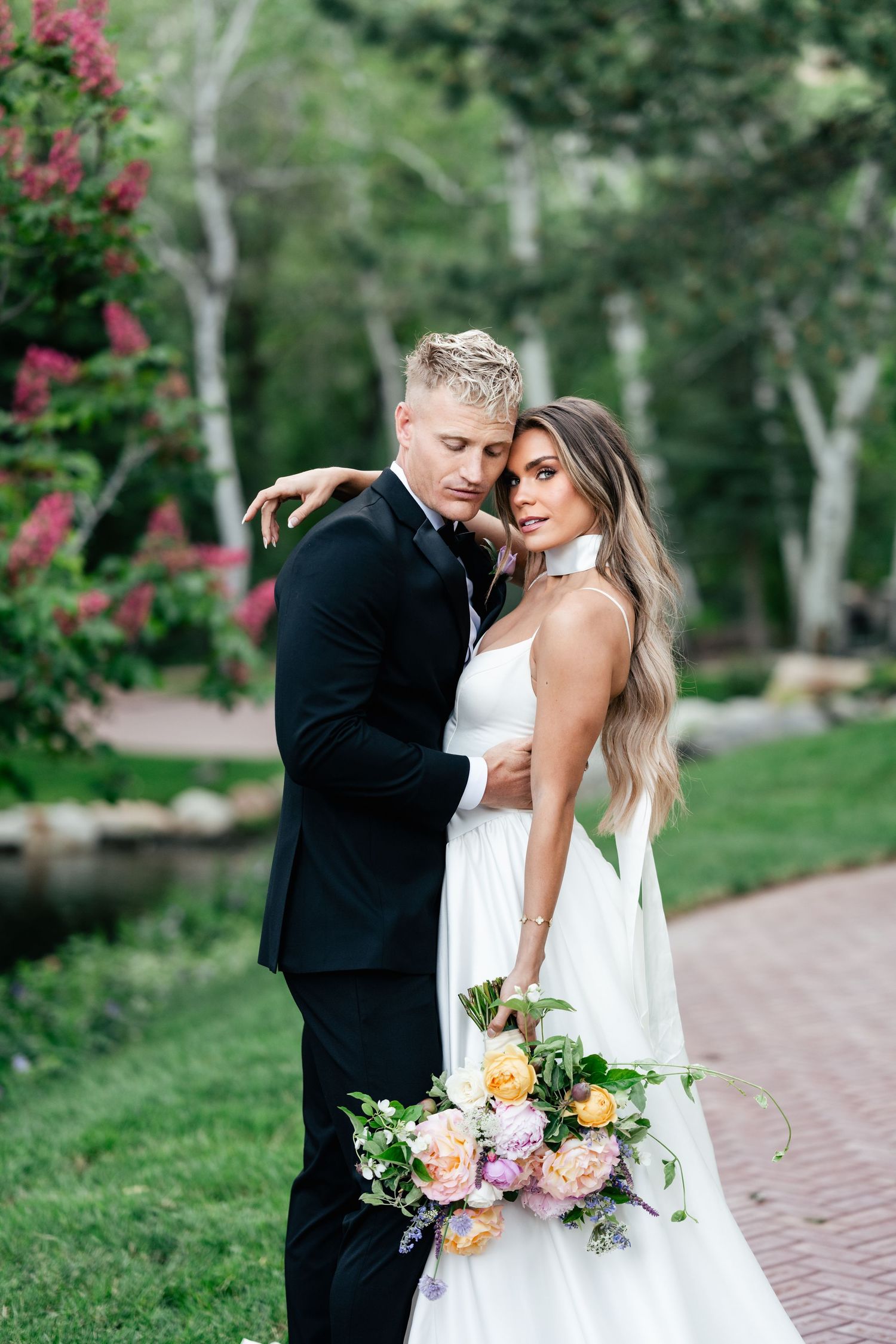 A couple in wedding attire embraces on a garden path surrounded by greenery while holding a colorful summer bouquet.