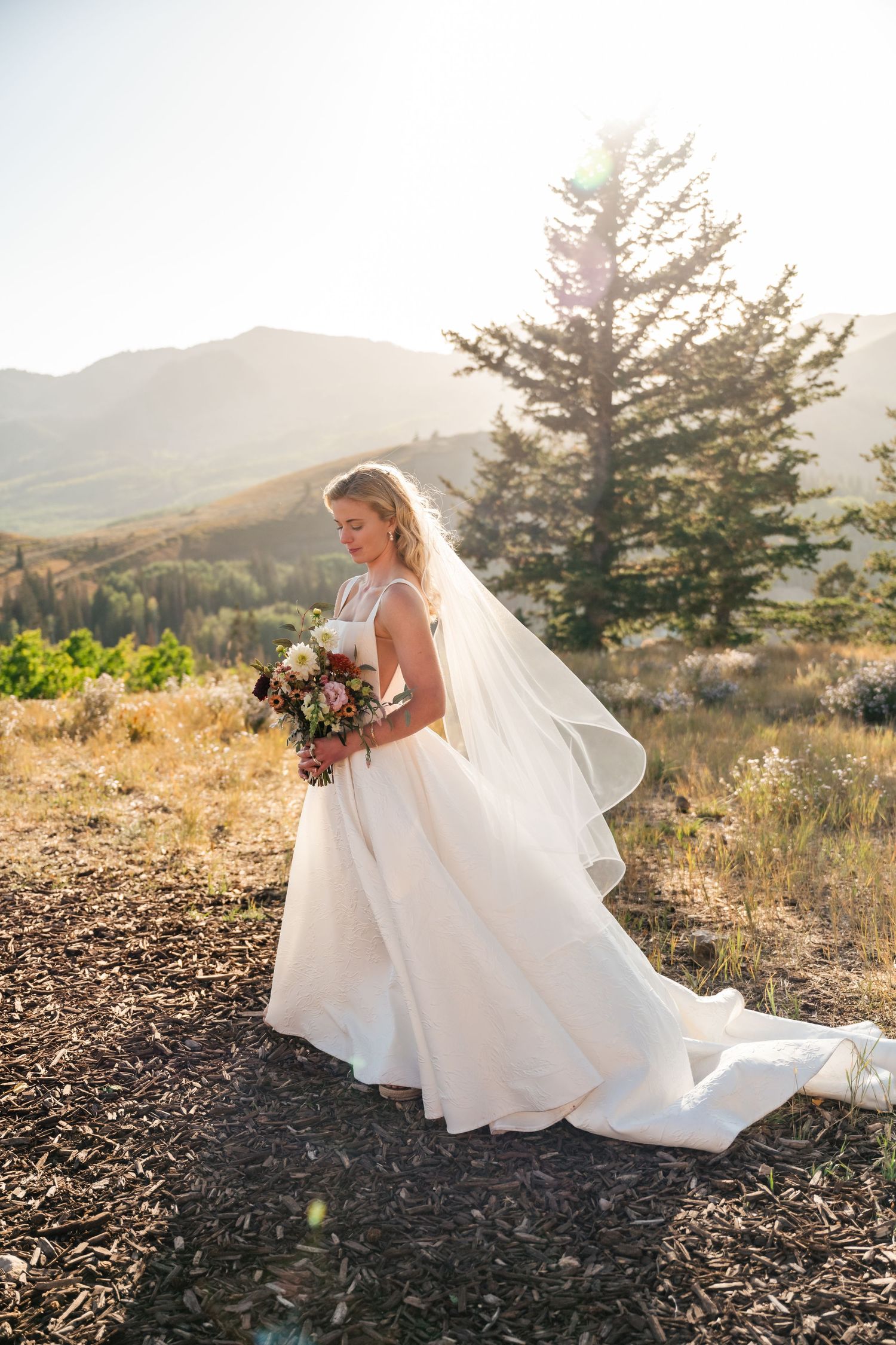 A bridal figure in a flowing white gown and veil holds a bouquet while standing on a scenic mountainside at sunset.