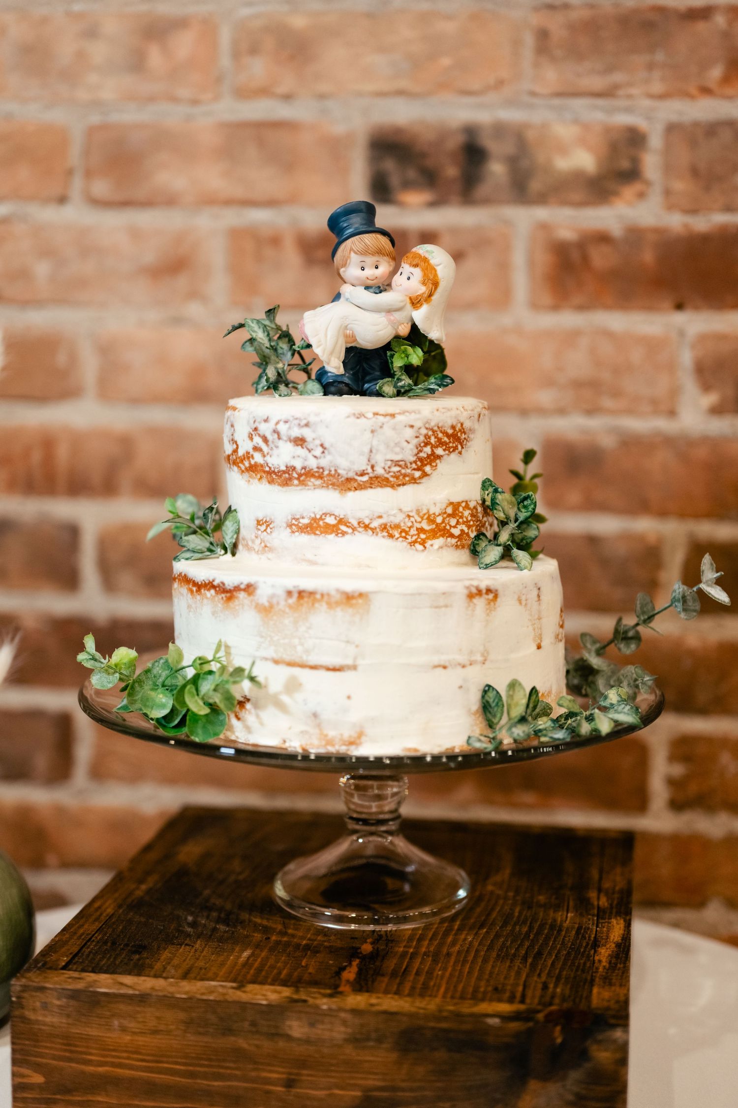 A rustic semi-naked wedding cake decorated with greenery and a whimsical garden gnome topper sits on a glass cake stand.