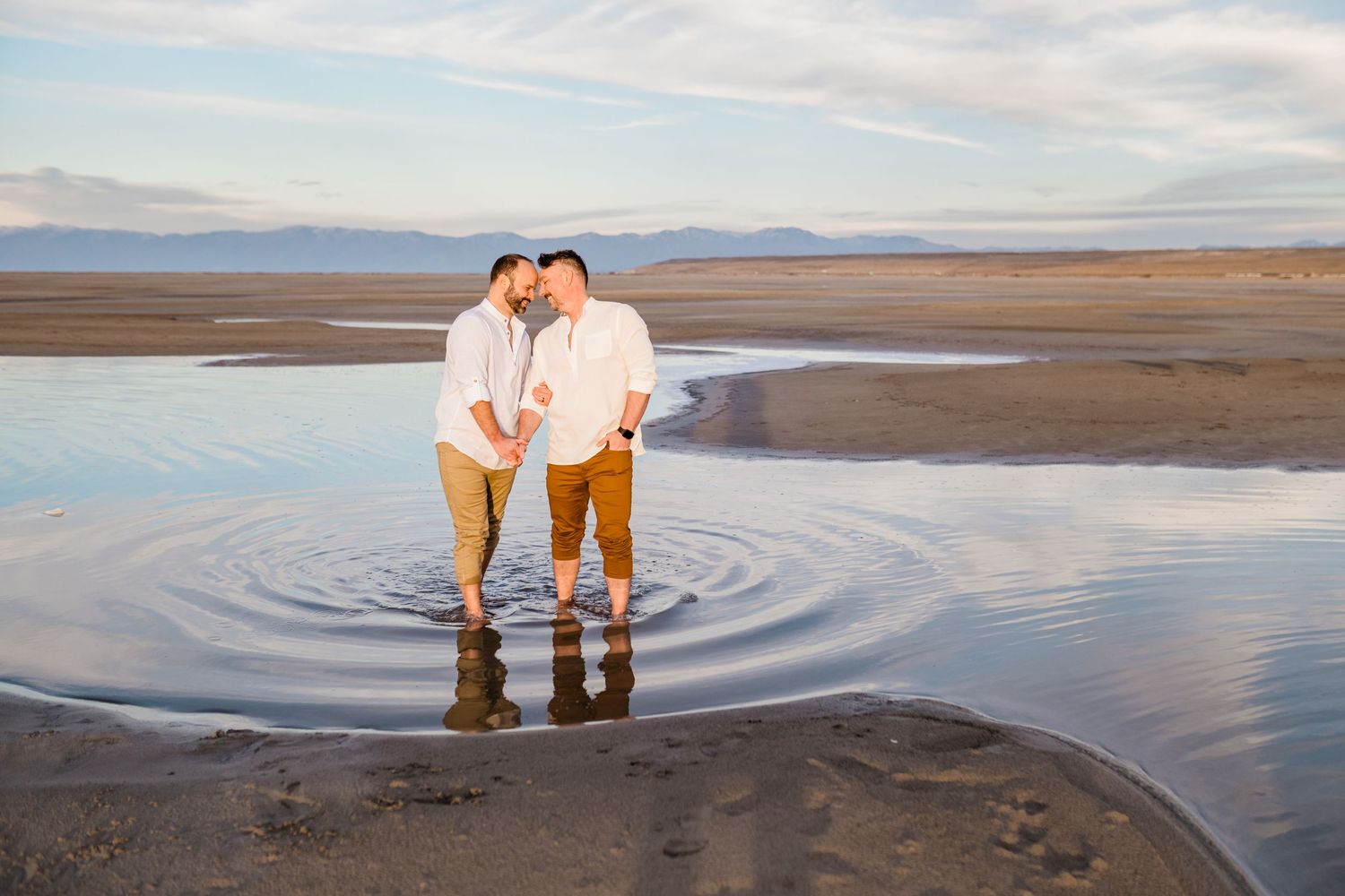 A couple stands in ankle-deep water at sunset on a beach with mountains in the distance.