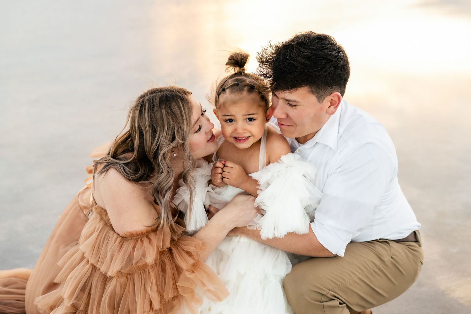 Parents embrace their child affectionately amidst the vast expanse of the flooded salt flats during golden hour.