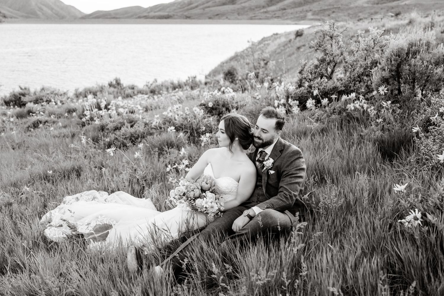 A couple sits together on a grassy hillside overlooking a scenic lake and mountain landscape in black and white.