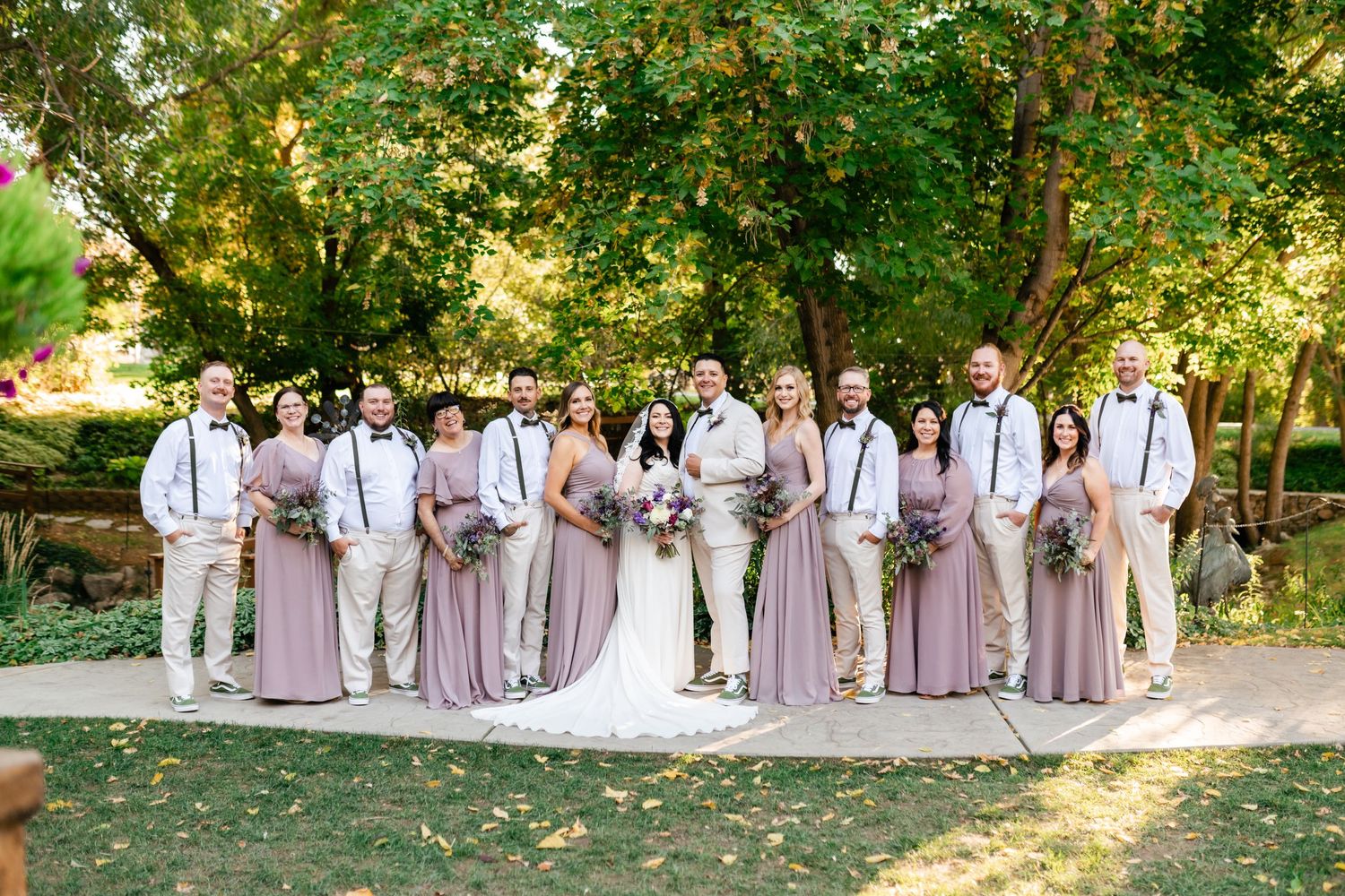 Wedding party wearing white shirts with suspenders and lavender dresses poses together in a garden setting.