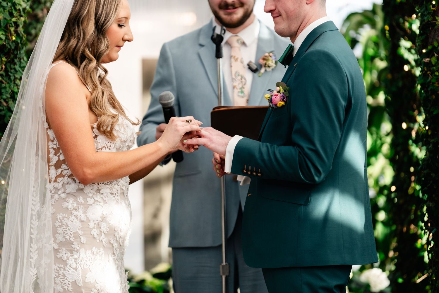 Wedding ceremony moment showing exchange of rings with officiant in gray suit and groom in emerald green jacket.