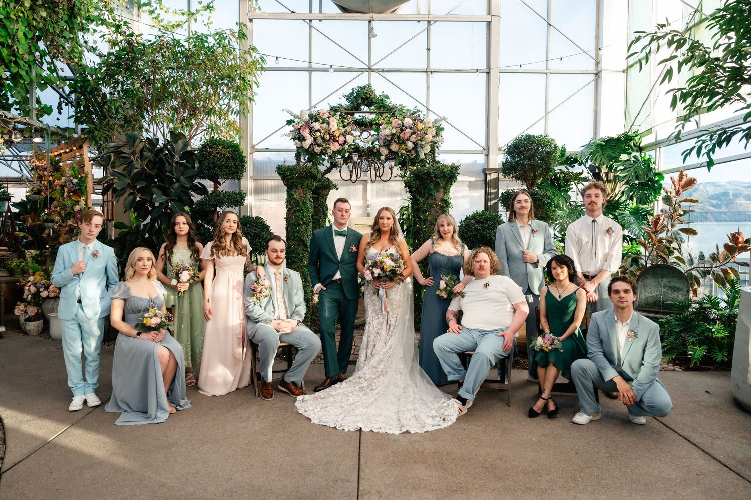 A wedding party poses in front of a decorative arch inside a bright greenhouse filled with lush greenery and natural light.