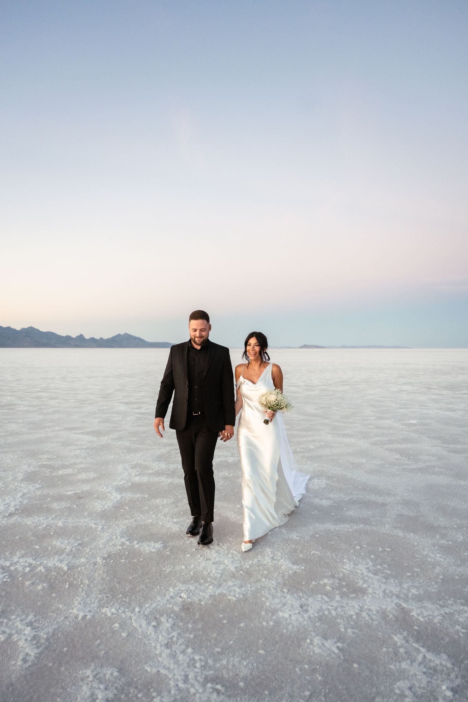 A couple walks across the expansive white salt flats during a wedding photo session at sunset.