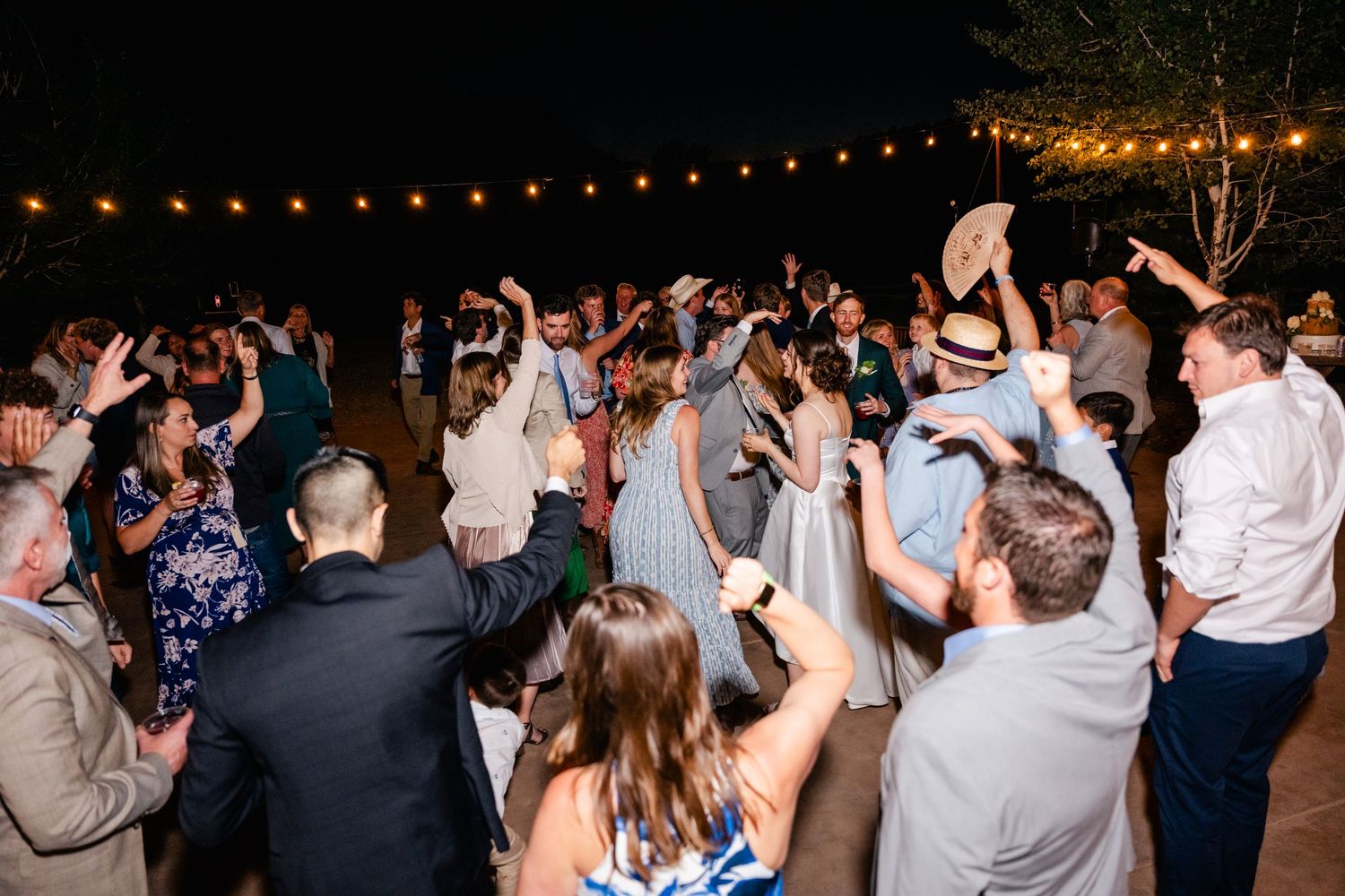 Large crowd dancing and celebrating at a nighttime outdoor wedding reception under string lights.