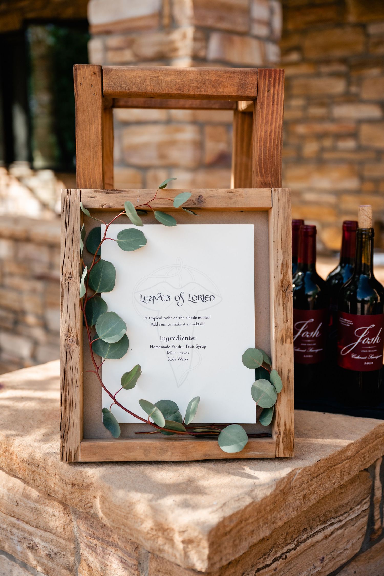 Rustic wooden frame displaying a wedding menu decorated with eucalyptus leaves beside wine bottles on stone surface.