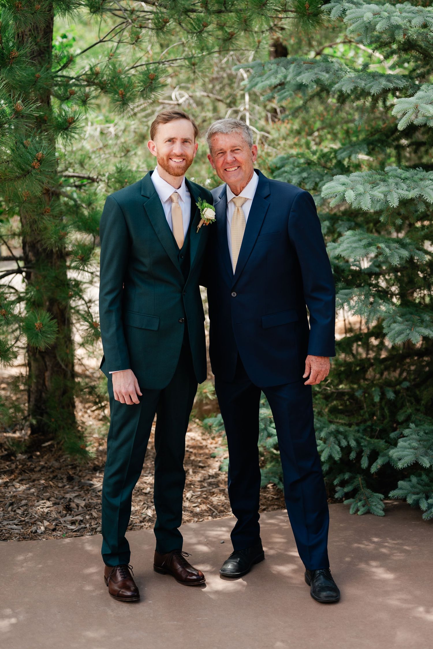 Two people in suits standing together outdoors with evergreen trees in the background.