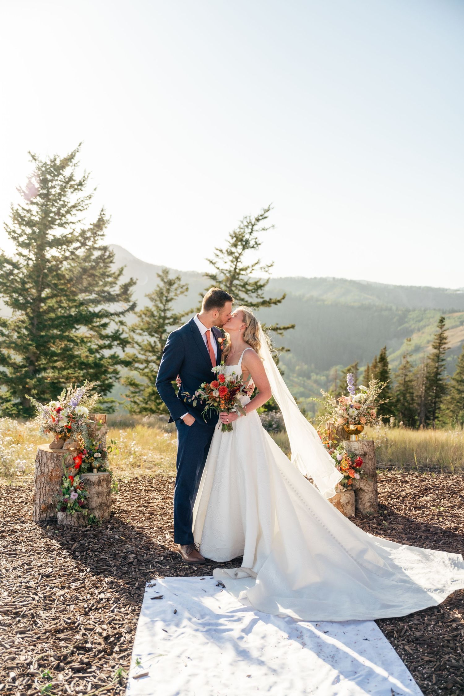 Newlyweds share a romantic moment against a mountain backdrop with evergreen trees and floral decor.