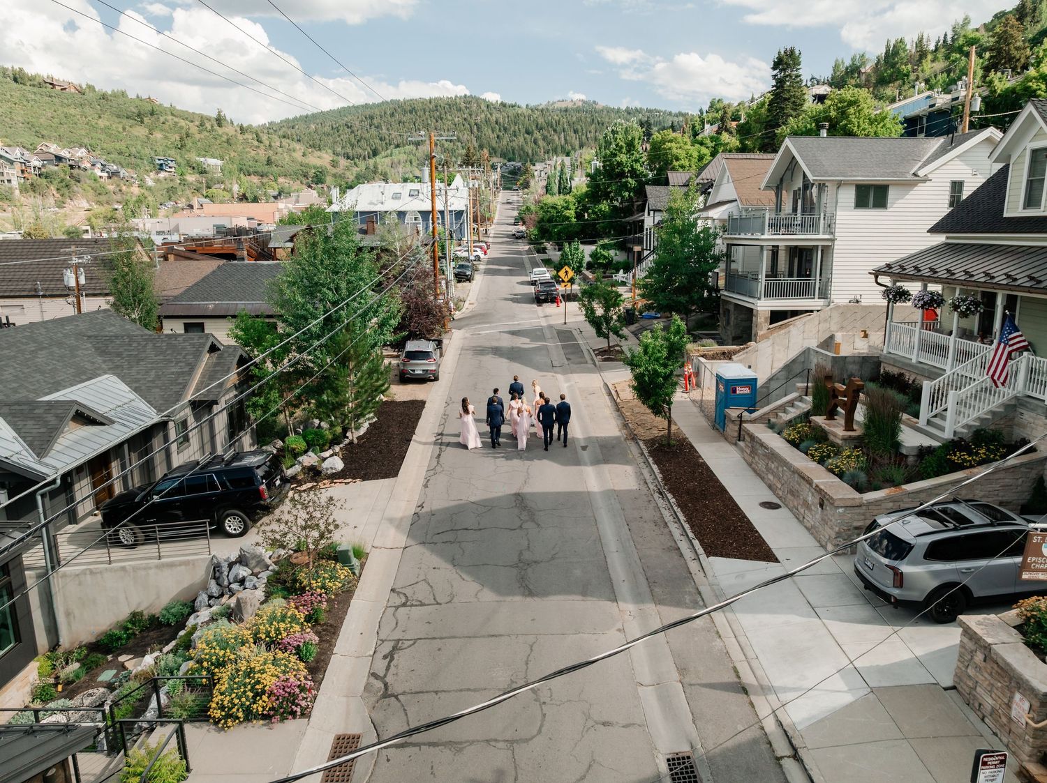 Aerial view of residential streets and buildings with parked cars and trees on a sunny day.