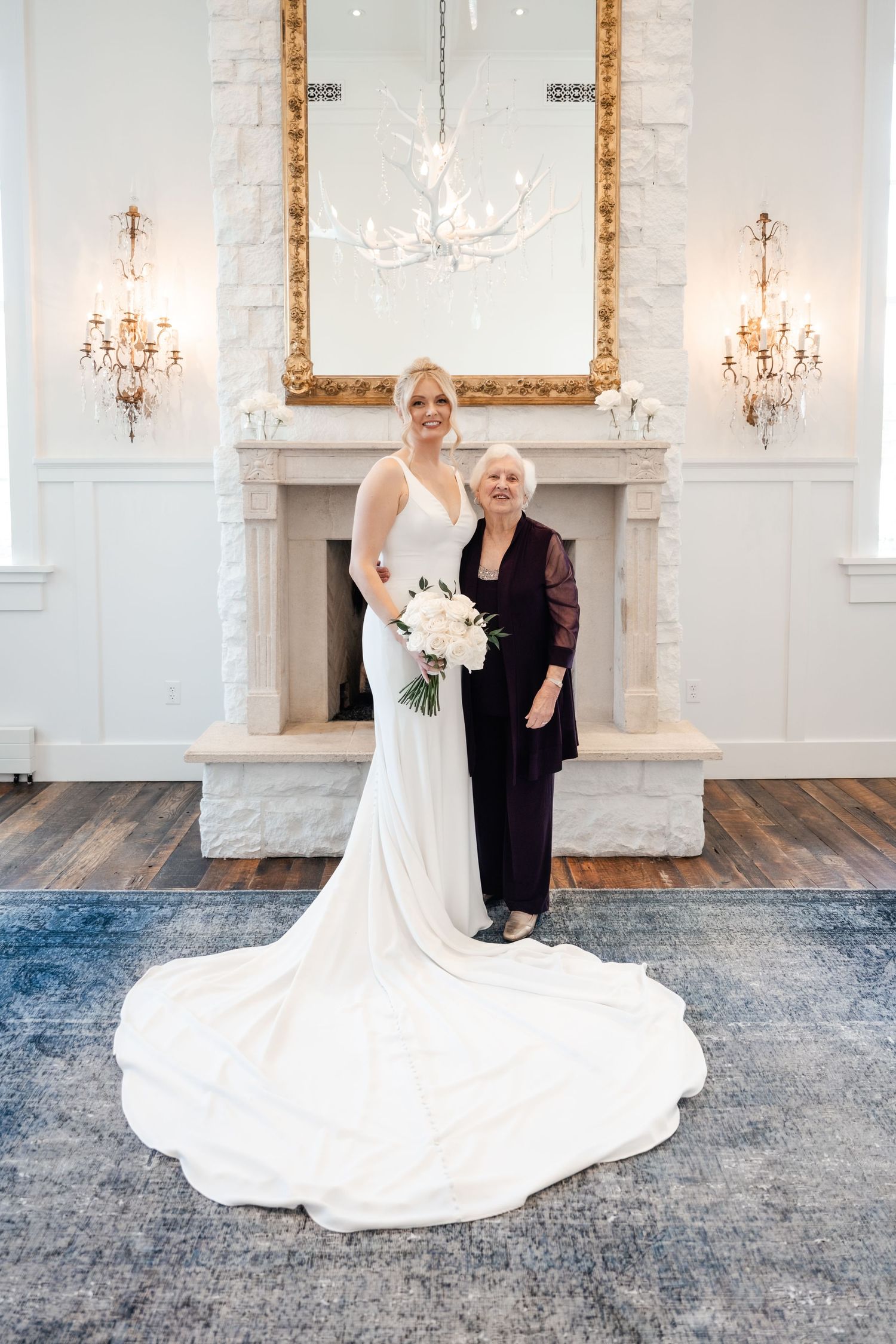 Elegant bride in flowing white gown with long train poses in grand room with ornate chandeliers and blue carpet.