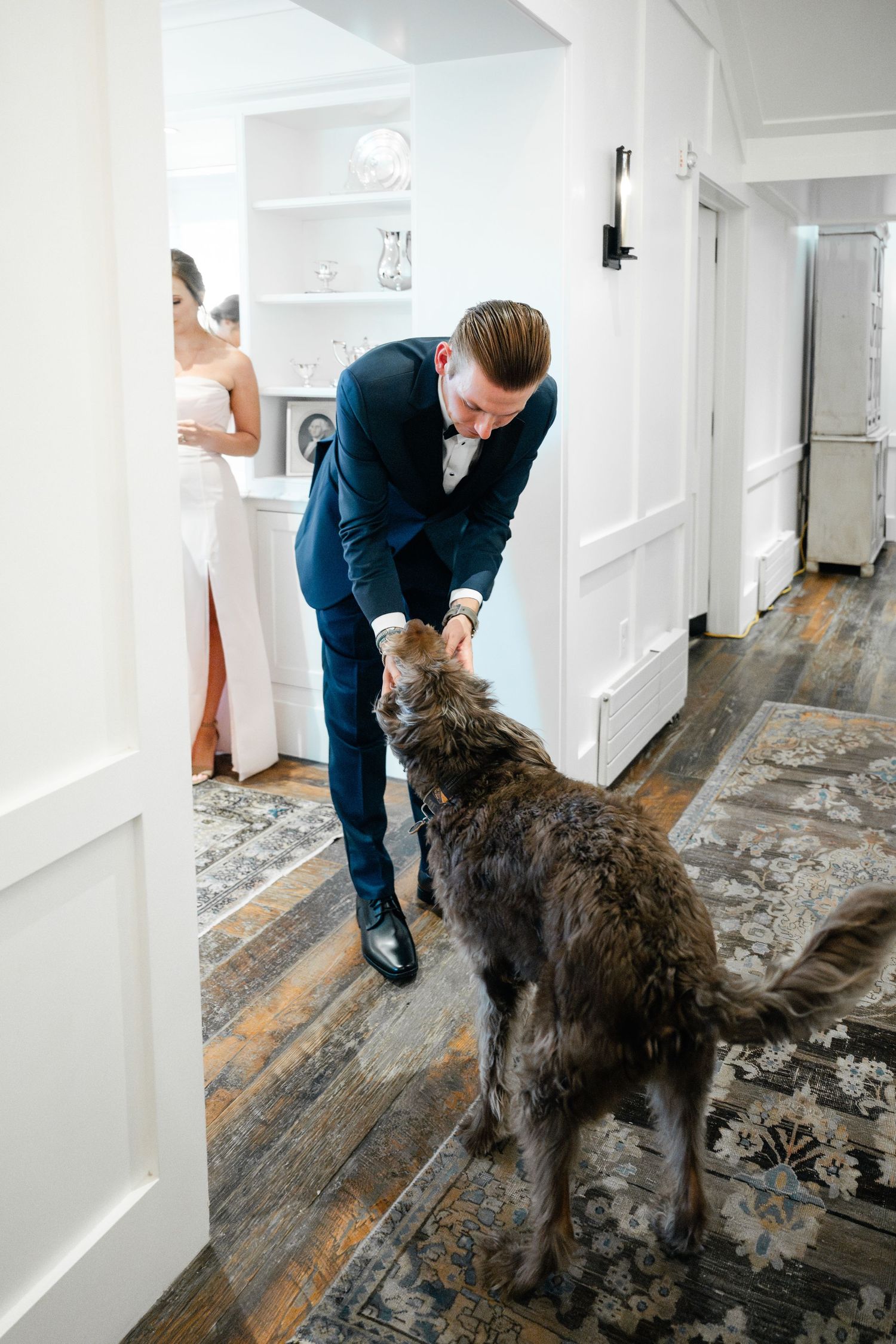 Man in navy suit petting large grey dog in white hallway.
