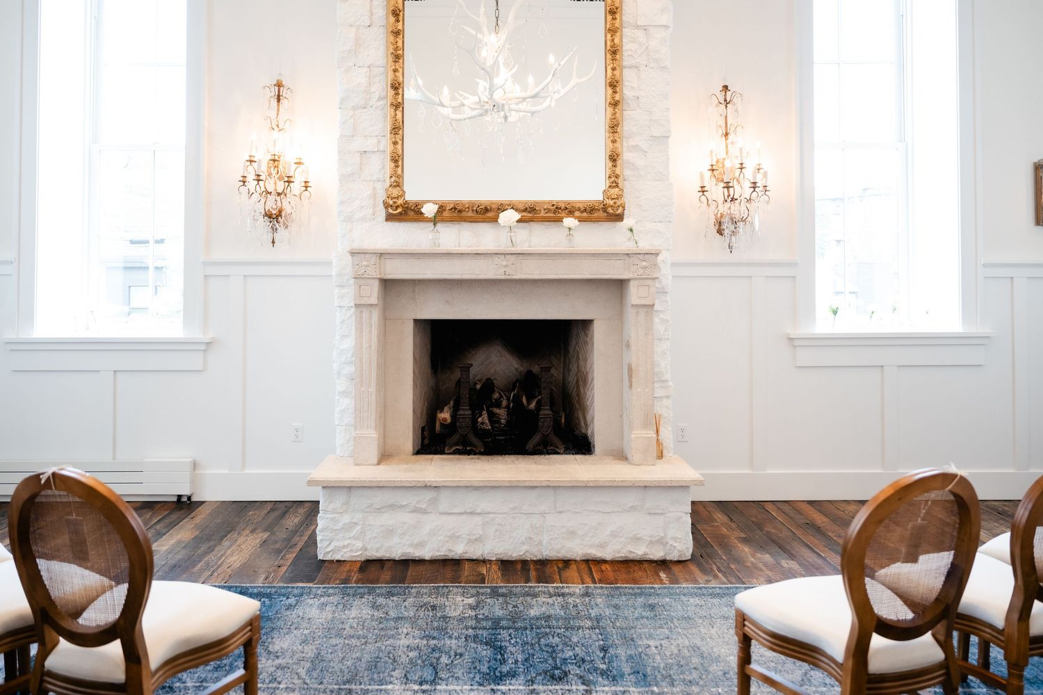 Elegant white room with marble fireplace, ornate gold mirror, crystal sconces and antique chairs.
