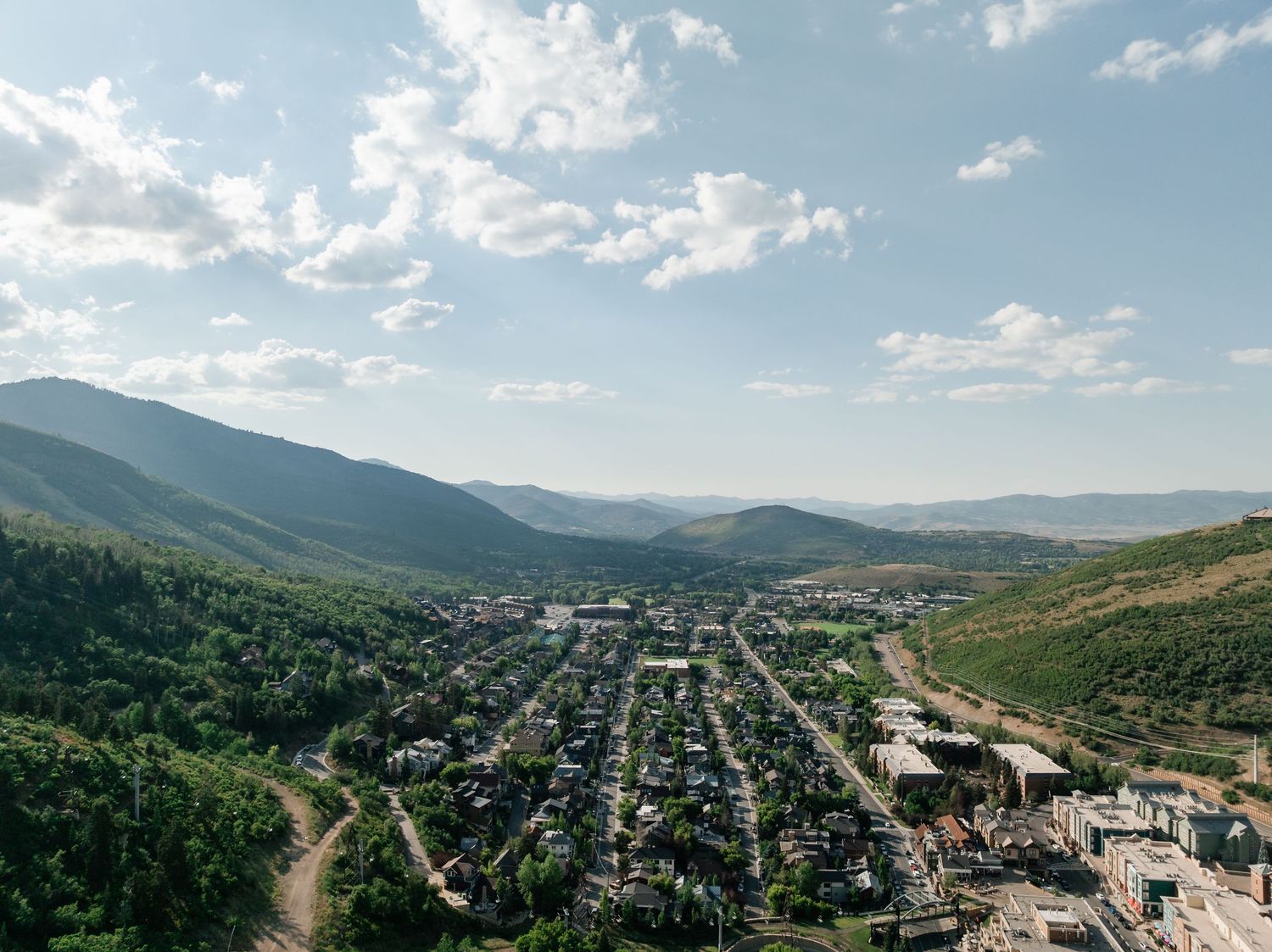 Aerial view of a small mountain town nestled in a valley with green hills and scattered clouds in the blue sky.