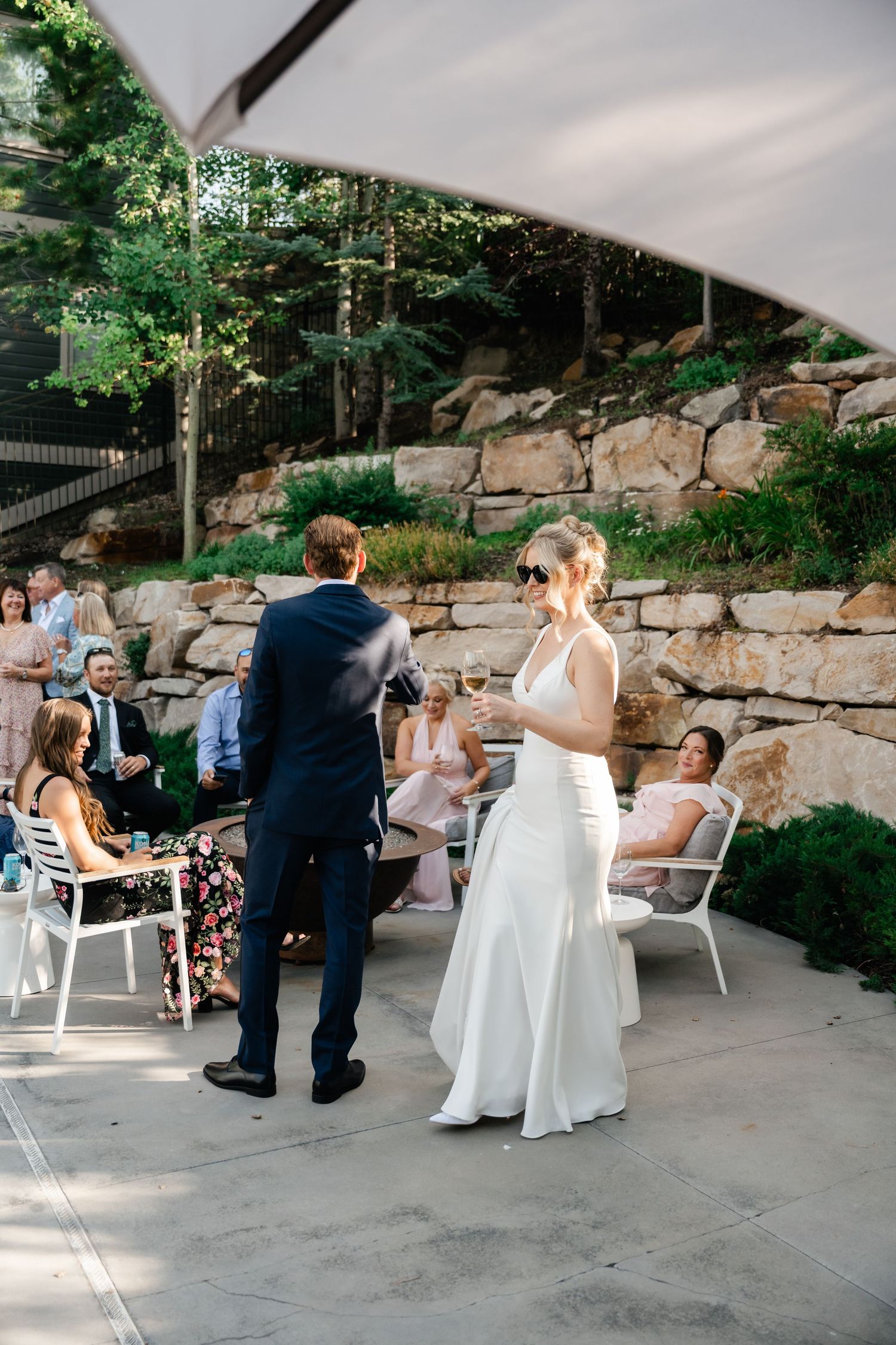 Wedding guests mingle and socialize on an outdoor patio with stone walls and white umbrellas.