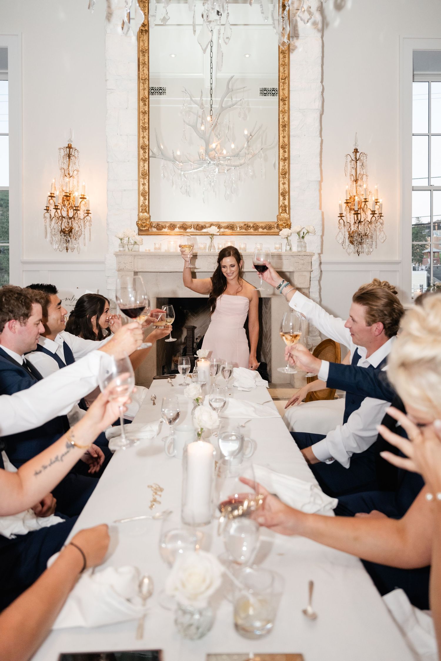 Wedding guests raise their glasses for a toast in an elegant white dining room with crystal chandeliers.