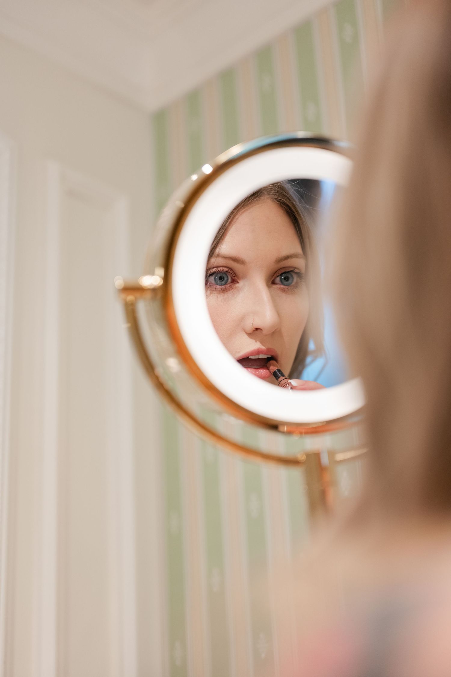 A circular illuminated makeup mirror reflects light in a bright room with striped wallpaper.