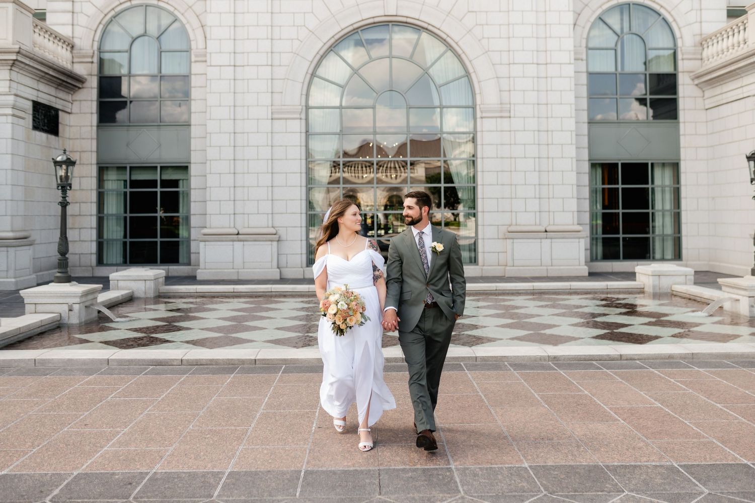 A couple walks together outside a grand building with arched windows and stone architecture on their wedding day.