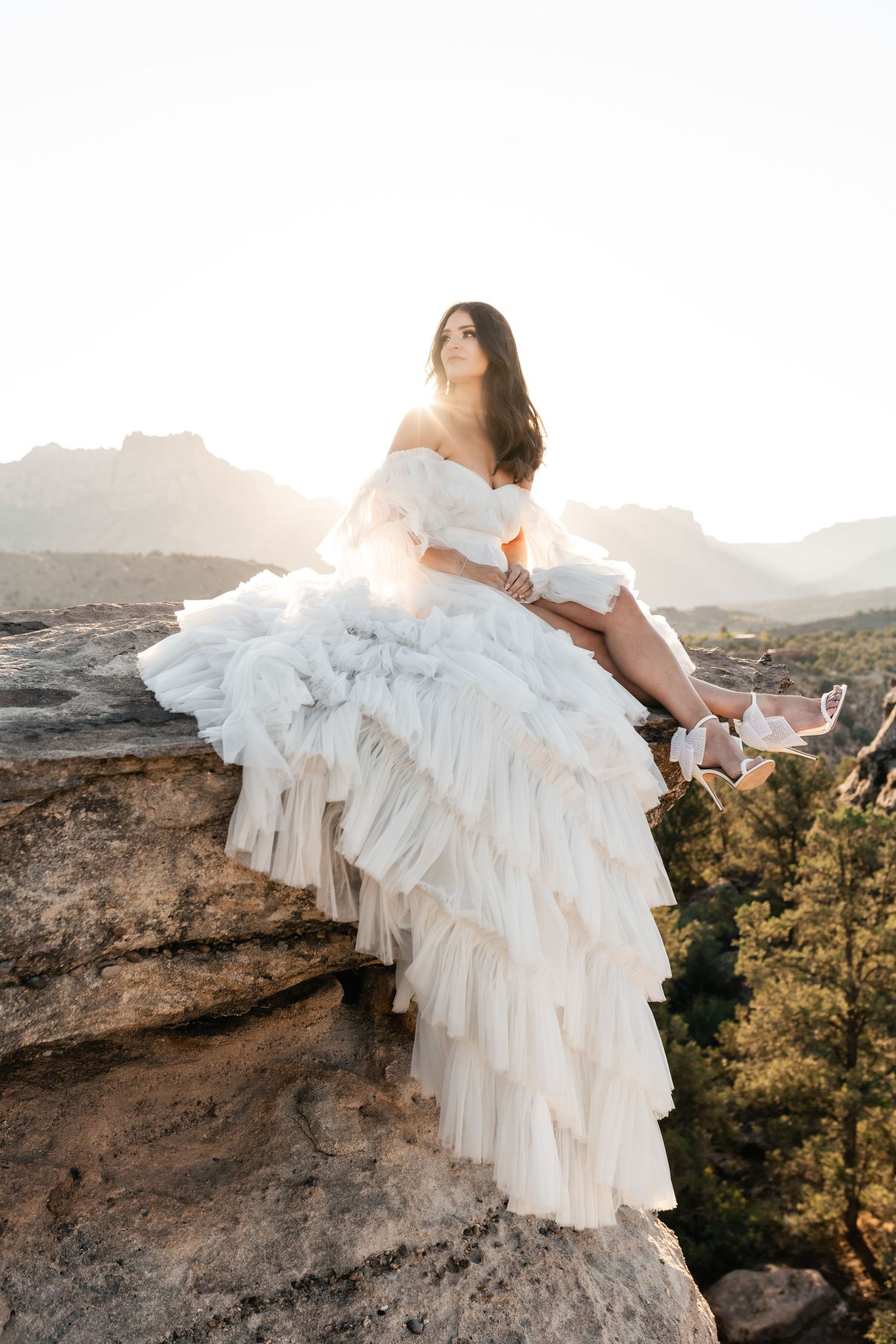 A bride in a flowing white ruffled gown sits gracefully on a rocky cliff edge at sunset in a mountainous landscape. Zion Bride.