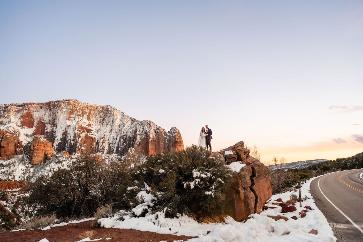 Snow-covered red rock formations and winding mountain roads create a dramatic winter landscape at sunset.
