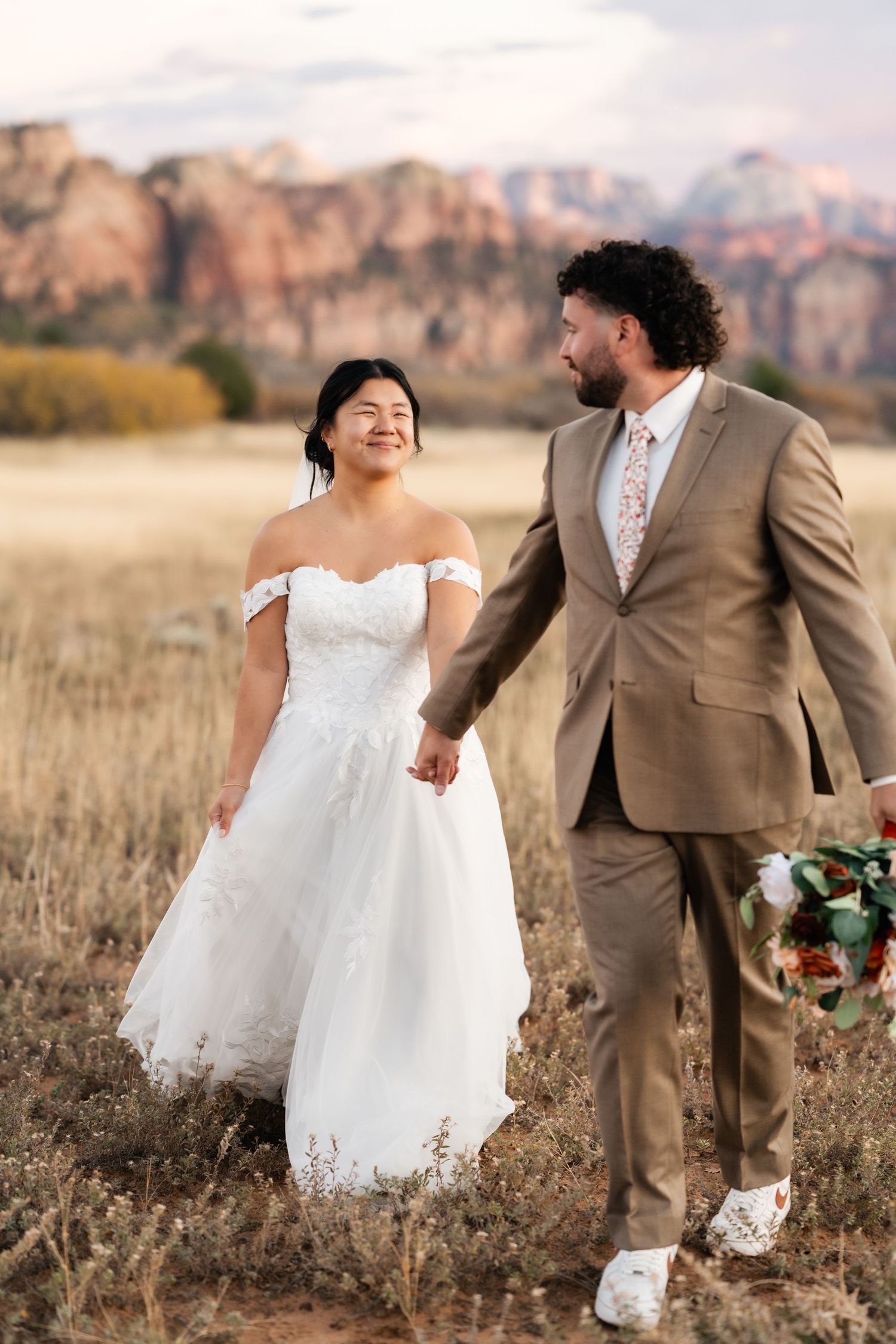 Newlyweds walk hand in hand through golden field with red rock mountains behind them.