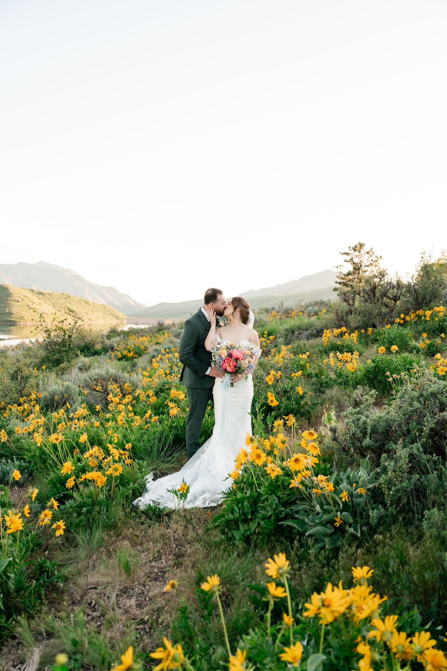 A romantic couple embraces in a field of vibrant yellow wildflowers with mountains. Park City Wedding Photographer, Coley Cook.