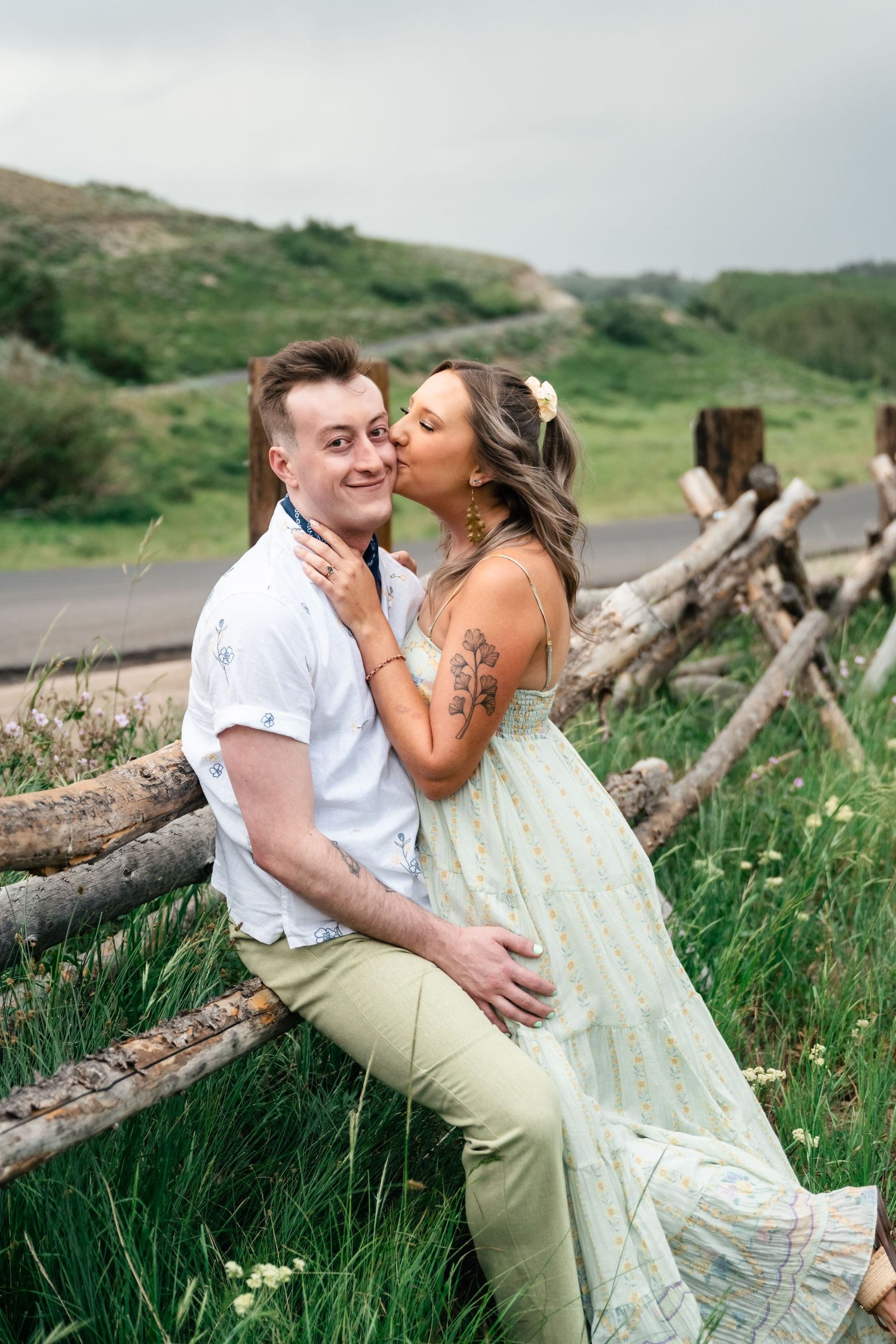 A couple shares intimate moments on a grassy hillside with rustic wooden fencing and mountain views in the background.