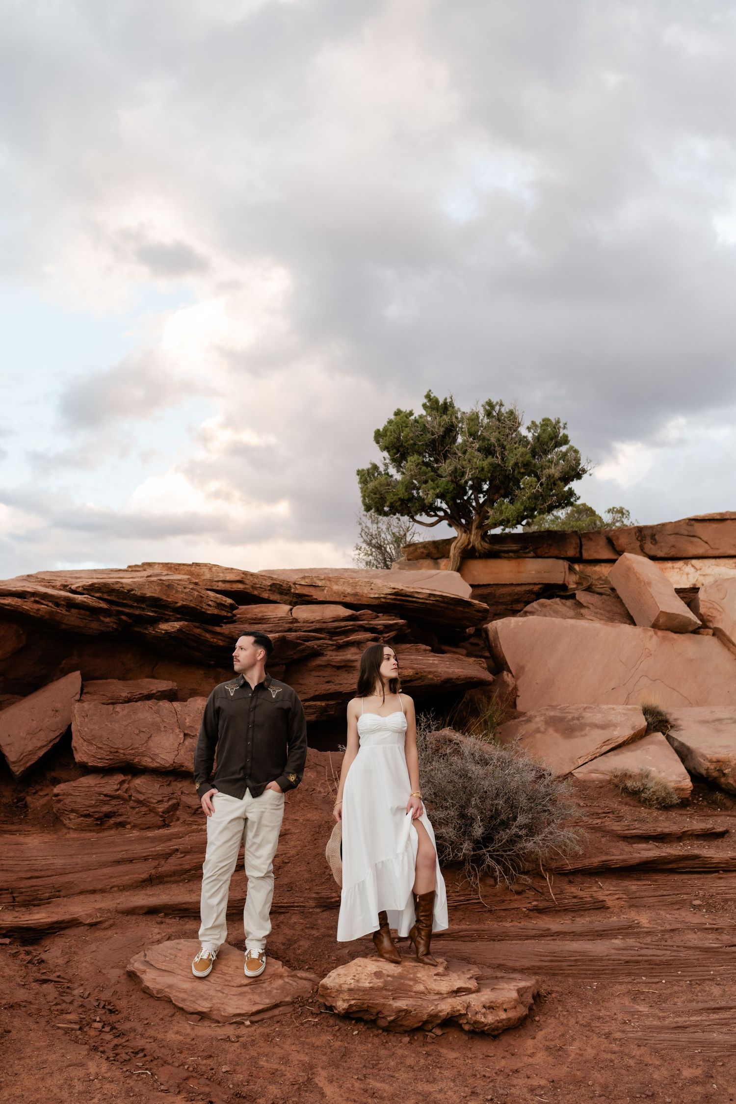 A couple stands together on red rock formations with a juniper tree and cloudy sky behind them.