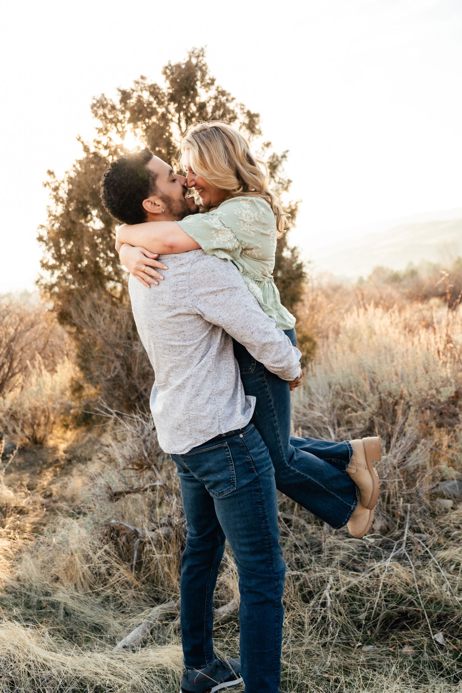 A romantic couple embraces in a sunlit field with golden grass and trees during sunset. Utah engagement photographer.