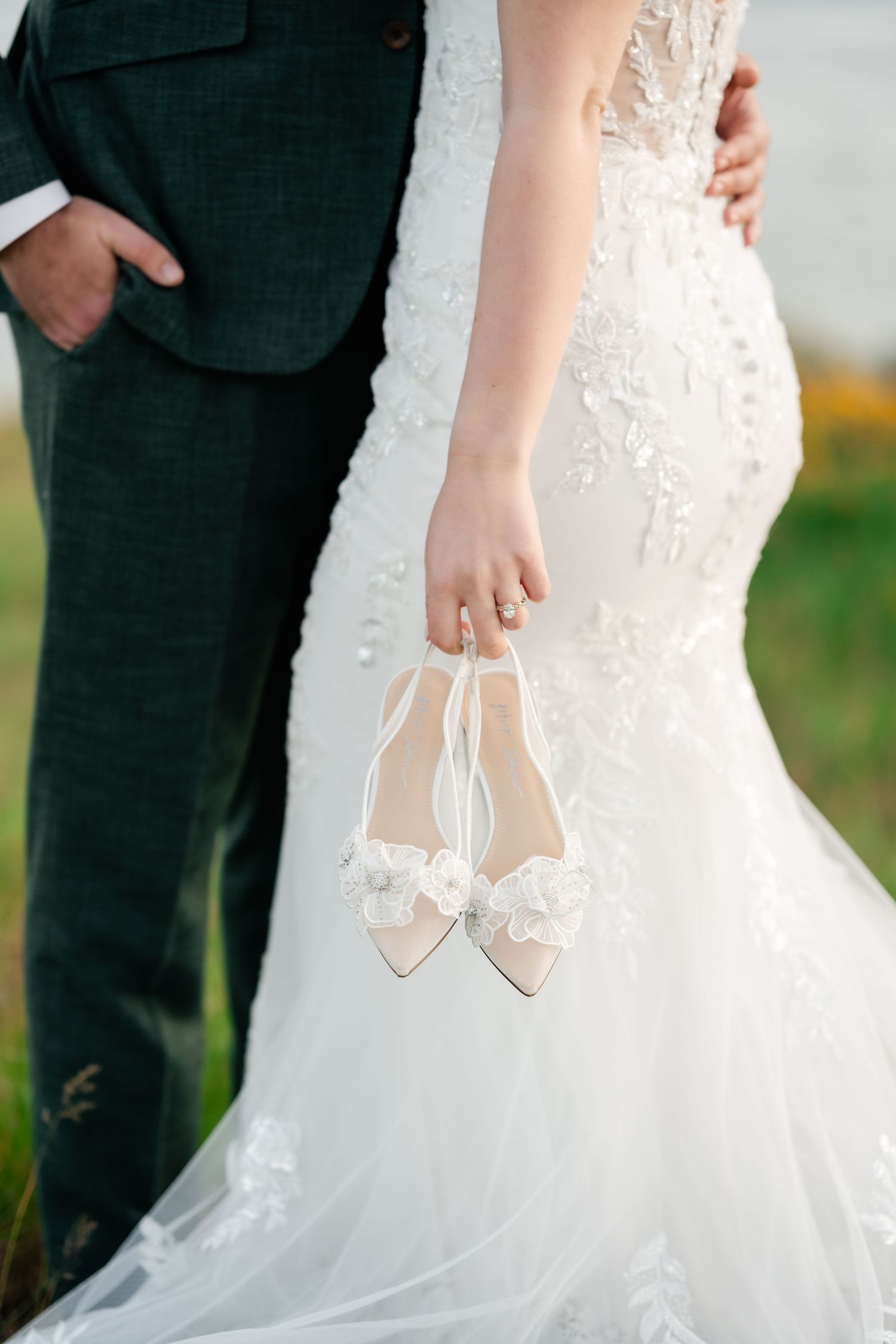 Close-up details of a bride's white wedding dress and shoes against a dark suit.