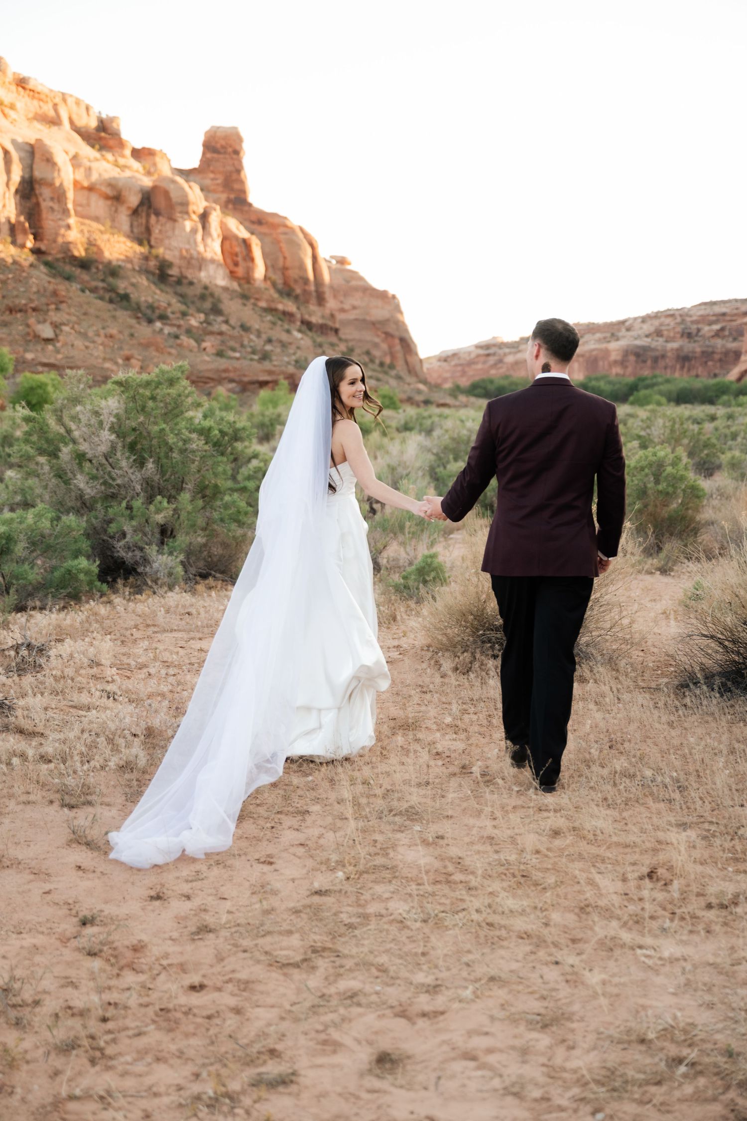 Bride looks back smiling while holding groom's hand walking through desert, her cathedral veil trailing, red rocks behind.