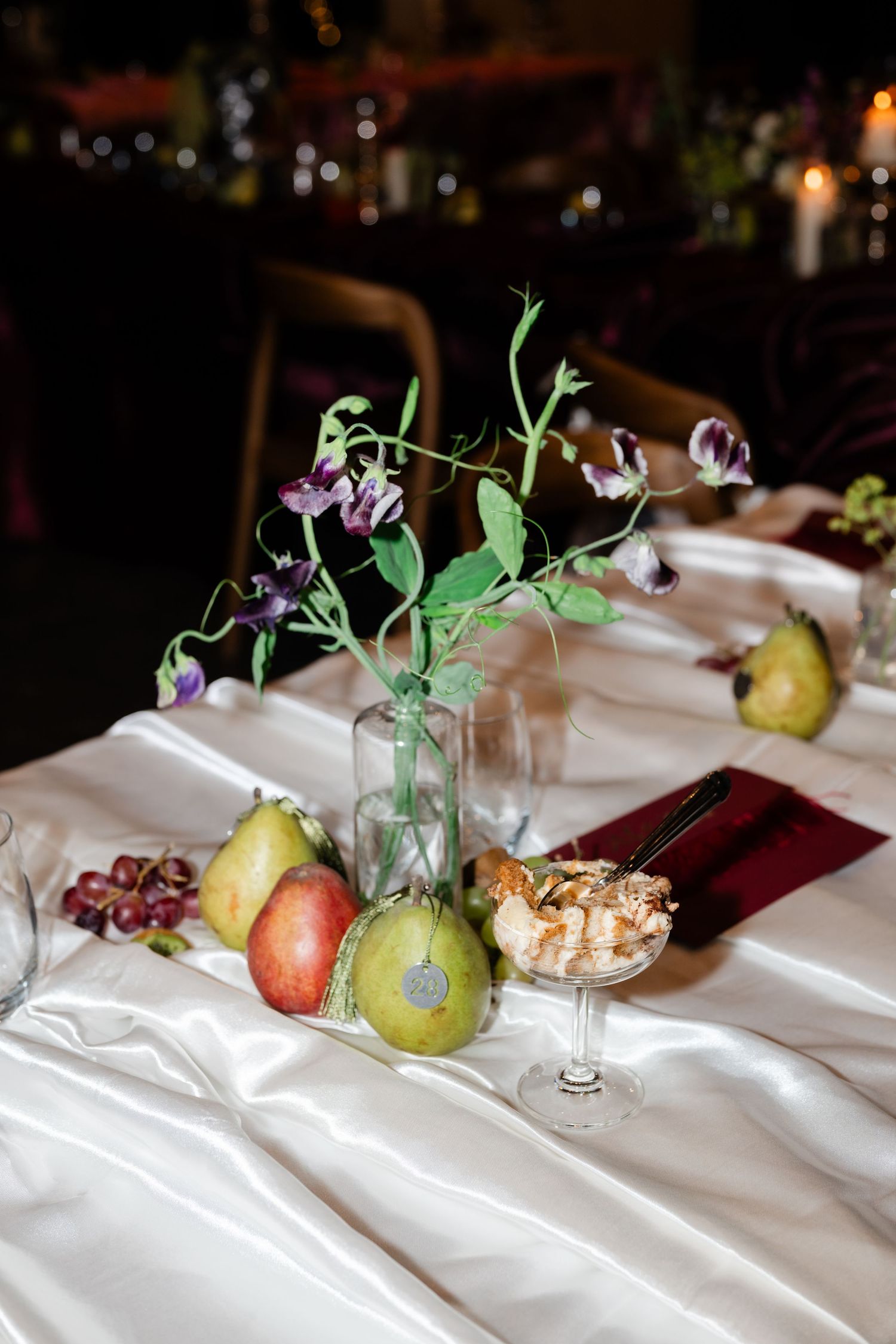 Elegant wedding table with white linen, glass vase with purple flowers, fresh pears, and a dessert cup centerpiece.