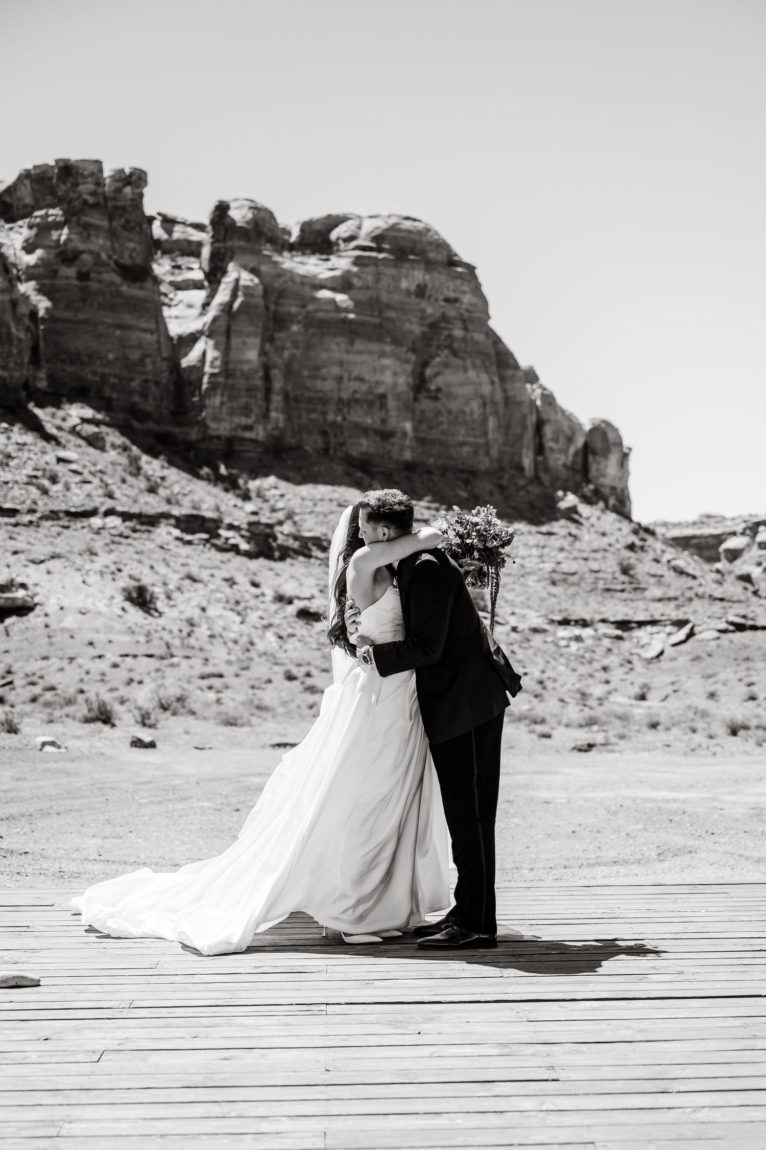 Black and white photo of a bride and groom embracing on a wooden deck with dramatic desert rock formations behind them.