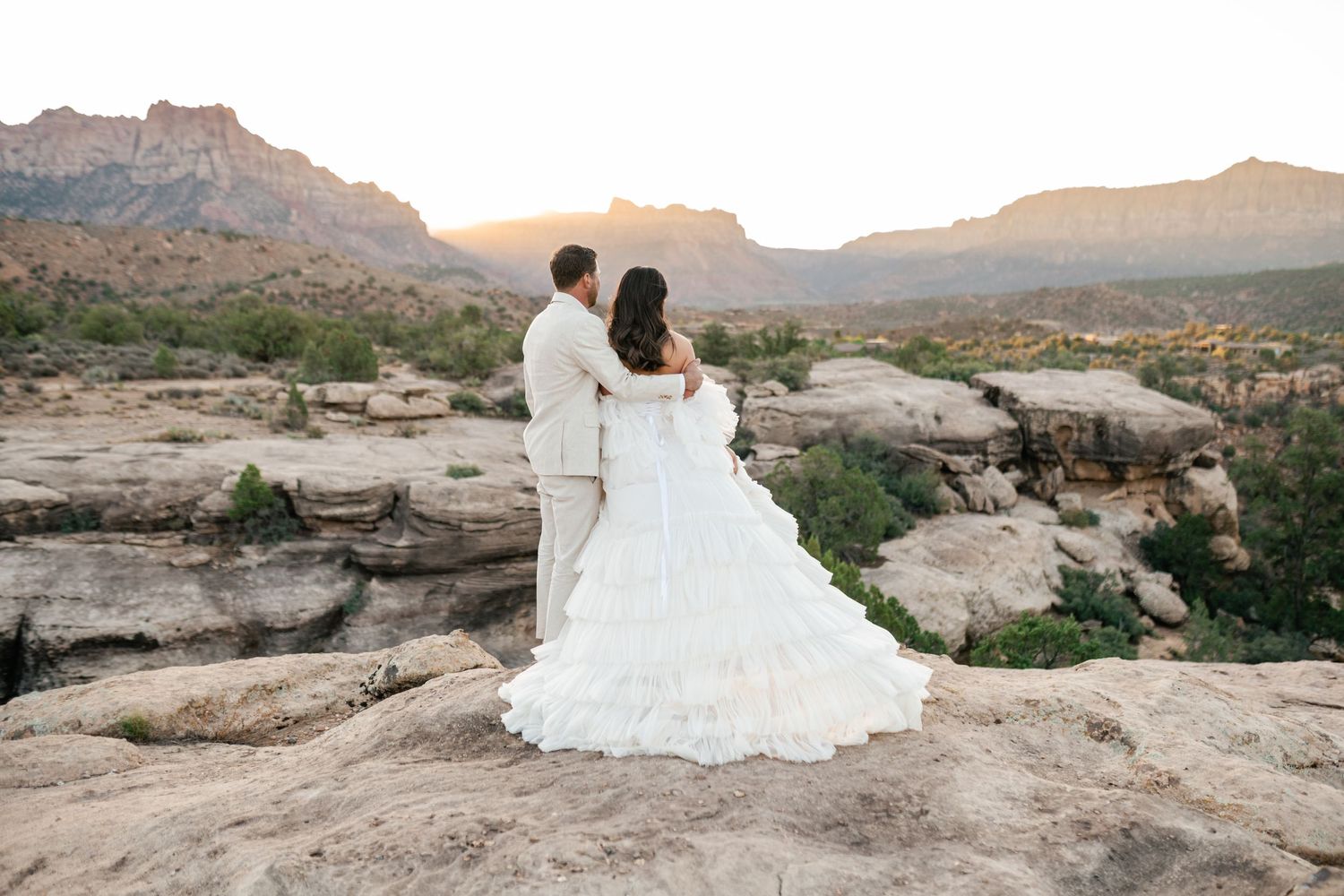 Zion Adventure Session. A couple in wedding attire stands atop a rocky mountain vista during sunset in a desert landscape.
