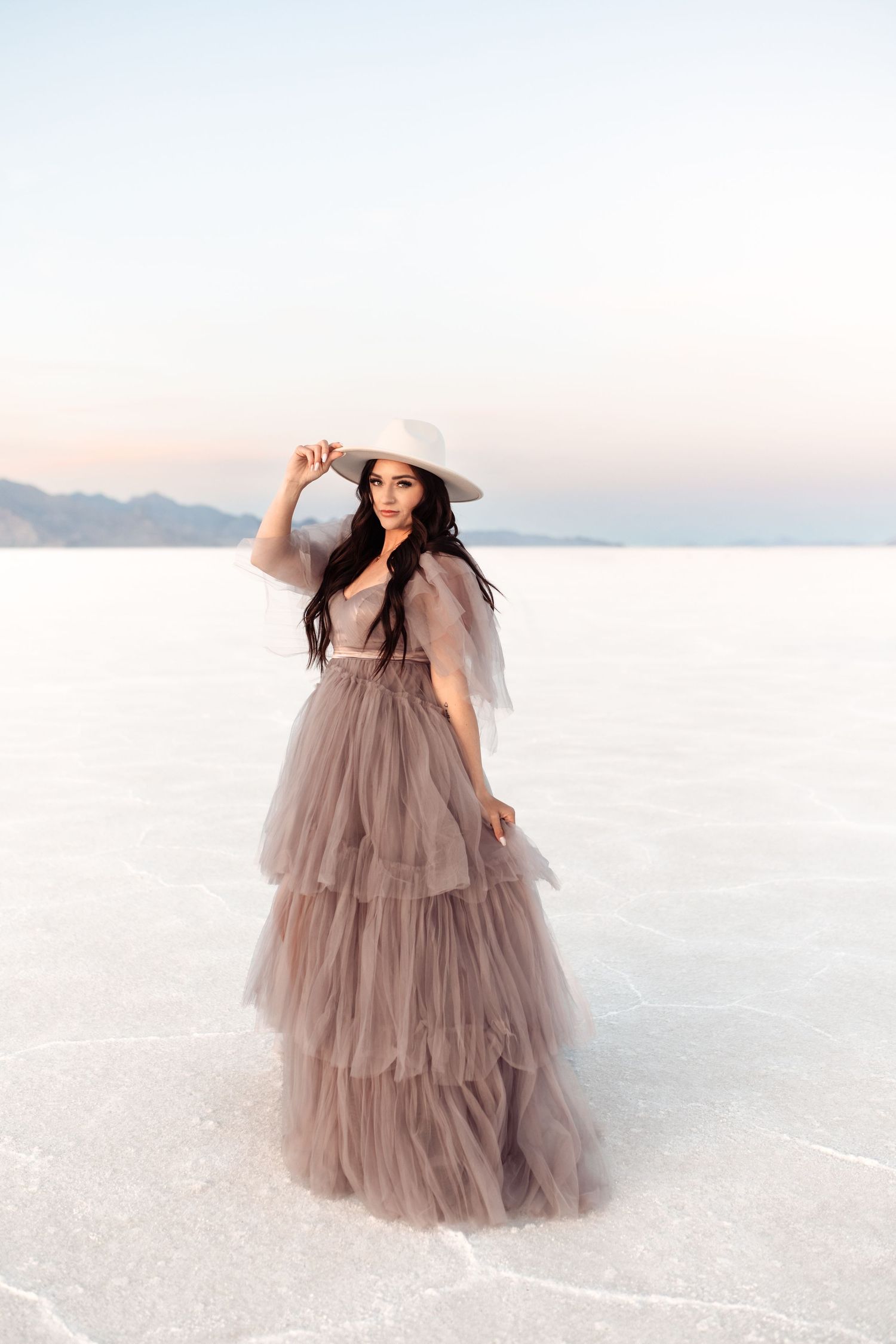 Utah Salt Flats Photographer. Woman in flowing mauve tiered tulle gown poses gracefully on salt flats under soft sky.