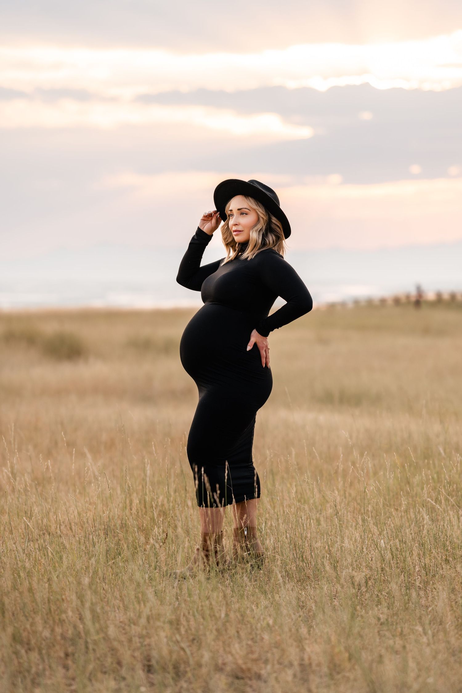 A figure in a black dress and hat stands expressively in a golden wheat field at sunset.