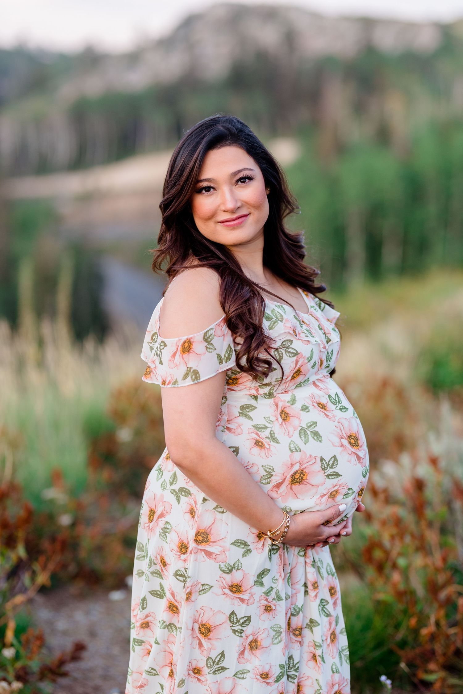 A woman in a floral dress poses outdoors in a natural setting with tall grass and greenery in the background.