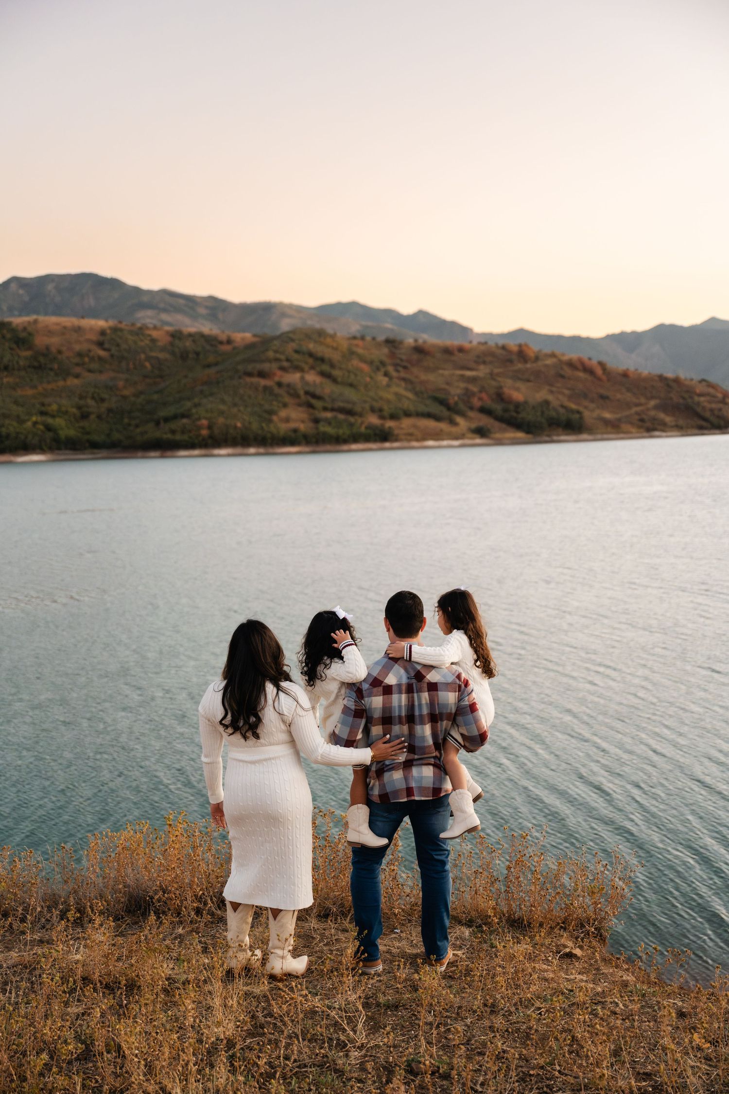 A family stands together at the edge of a scenic lake during sunset with mountains in the background.