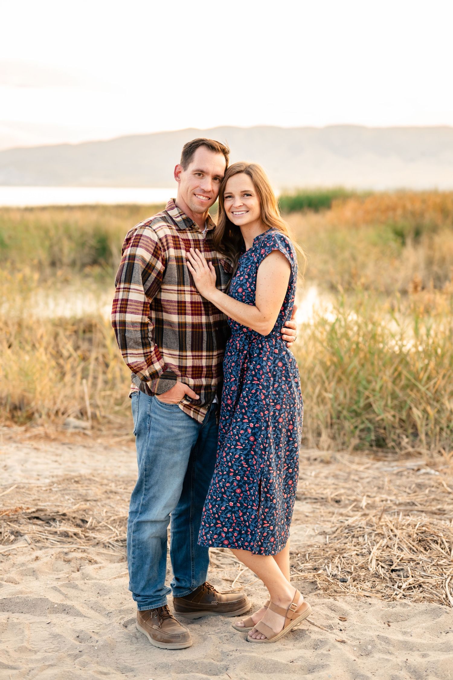 A couple poses together in a golden field at sunset wearing a plaid shirt and blue floral dress.