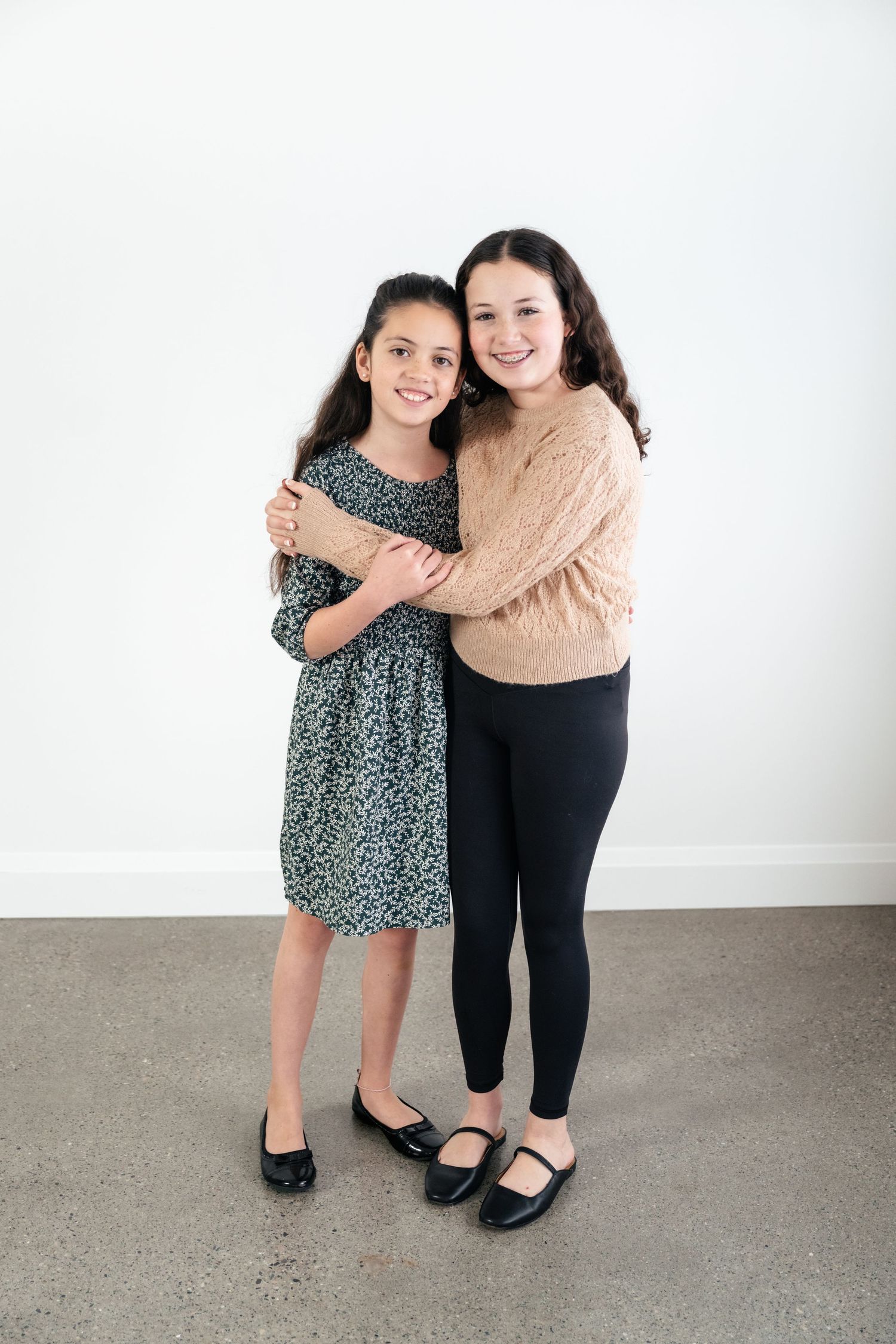 Two people embrace while standing against a white wall, wearing casual attire including a beige sweater and floral dress.