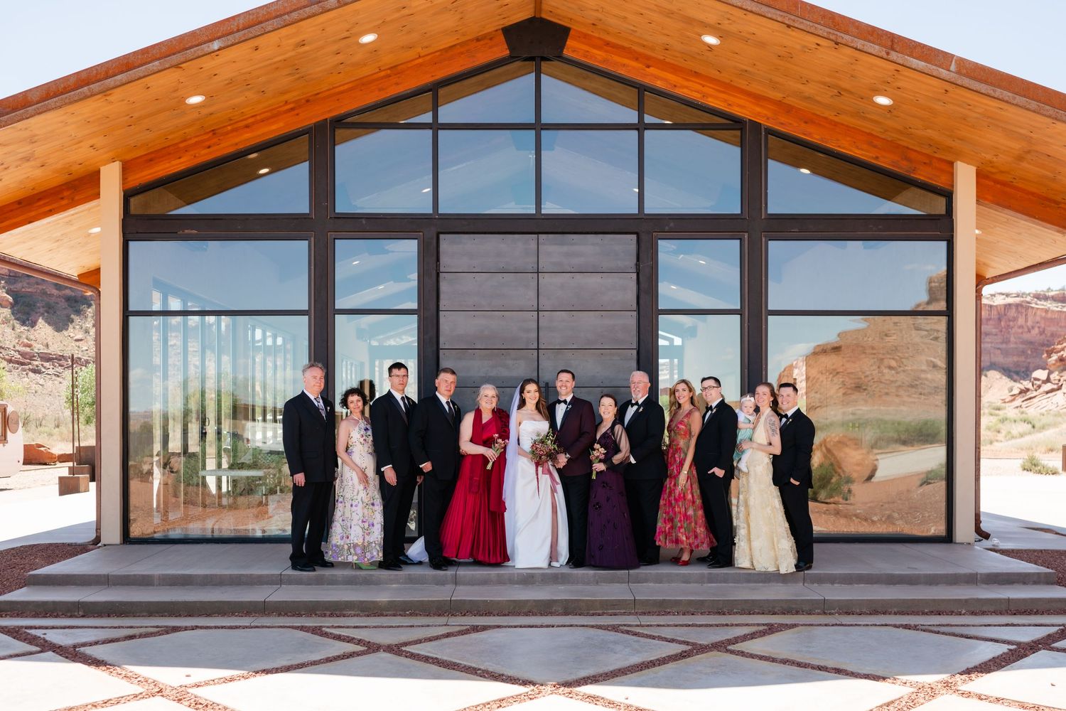 Wedding party posing in front of the red earth venue with large glass windows and desert landscape backdrop.