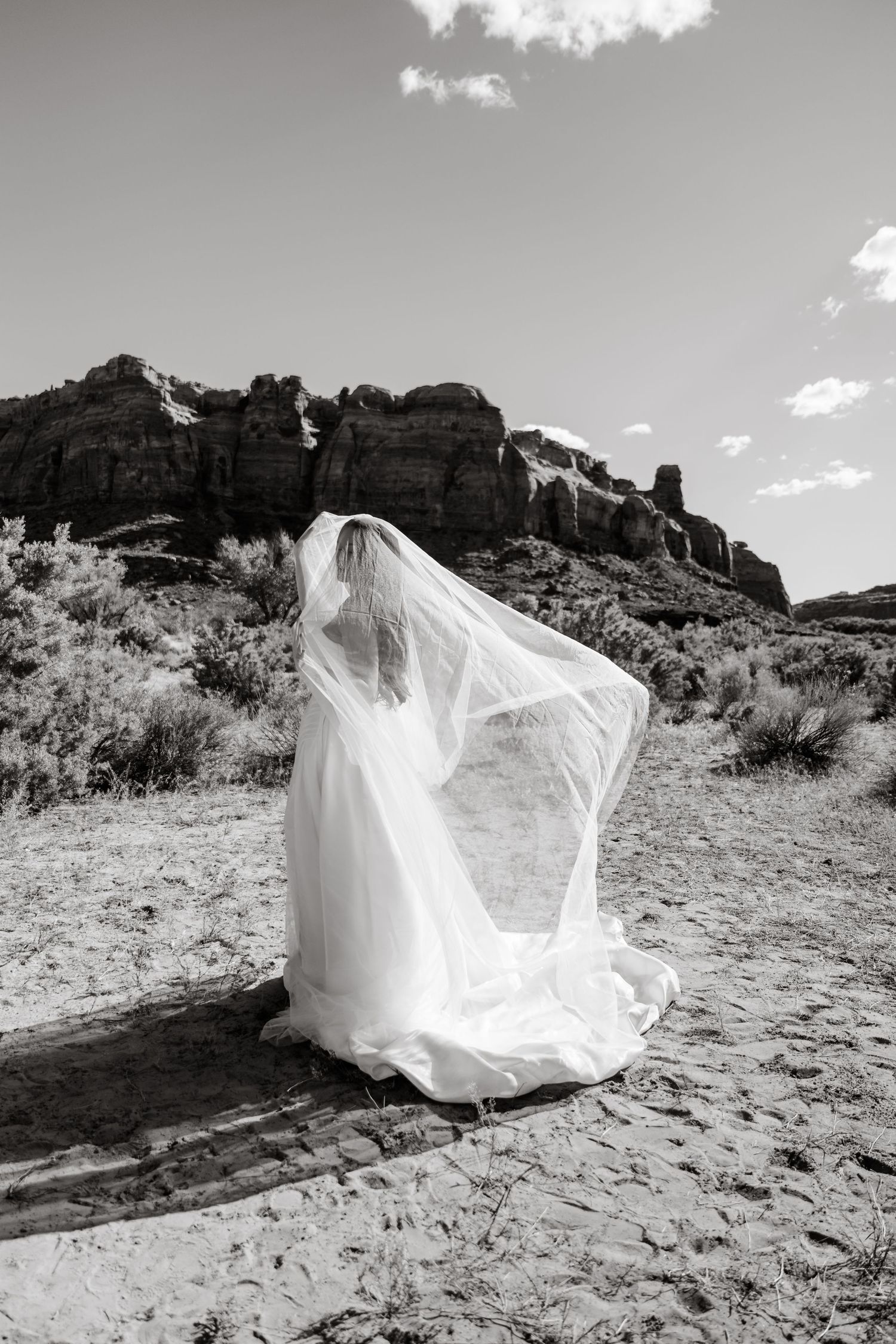Black and white photo of bride in flowing veil walking through red rock desert landscape.