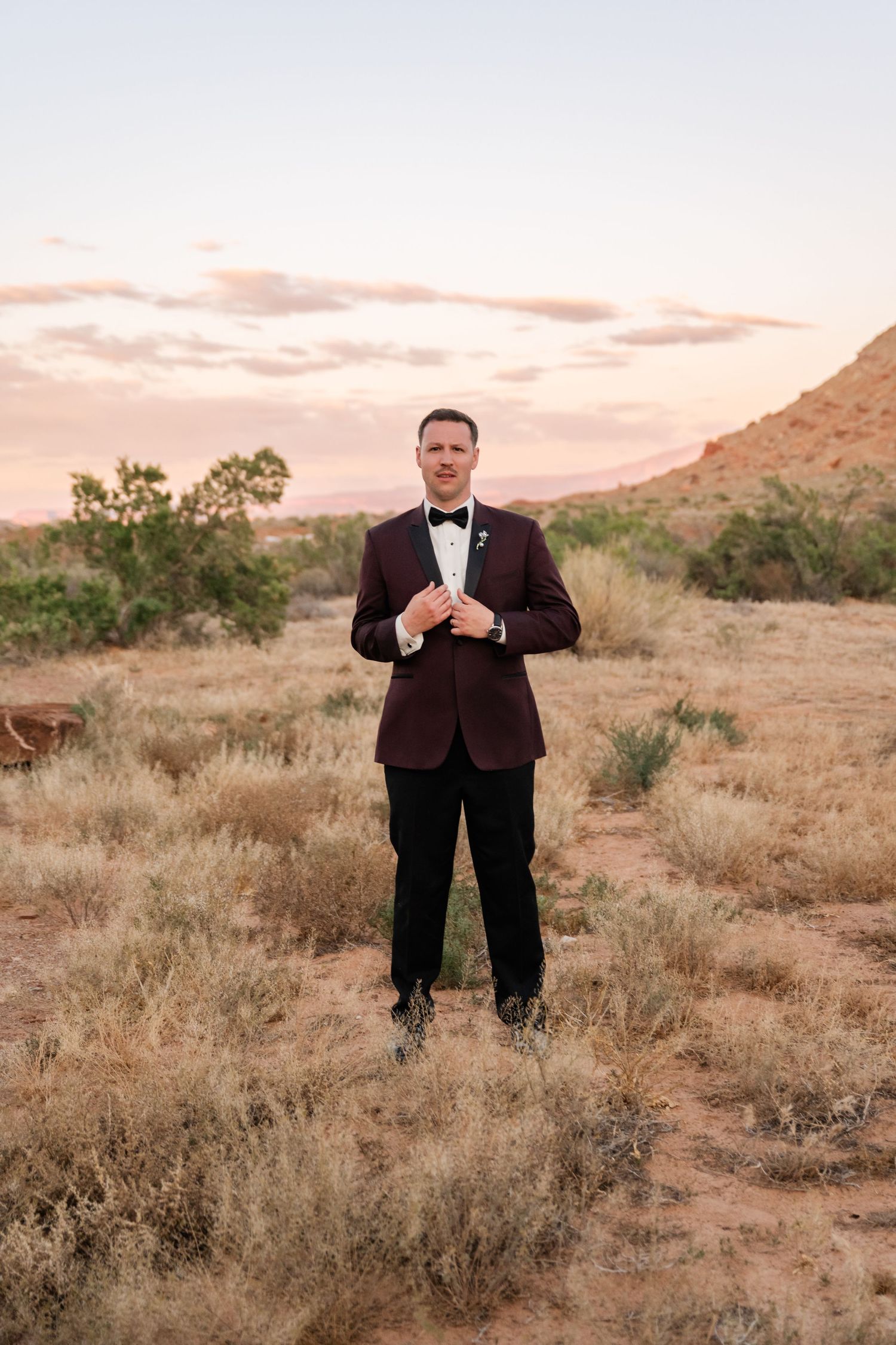 Groom in burgundy tuxedo jacket with black bow tie standing in desert landscape at sunset.