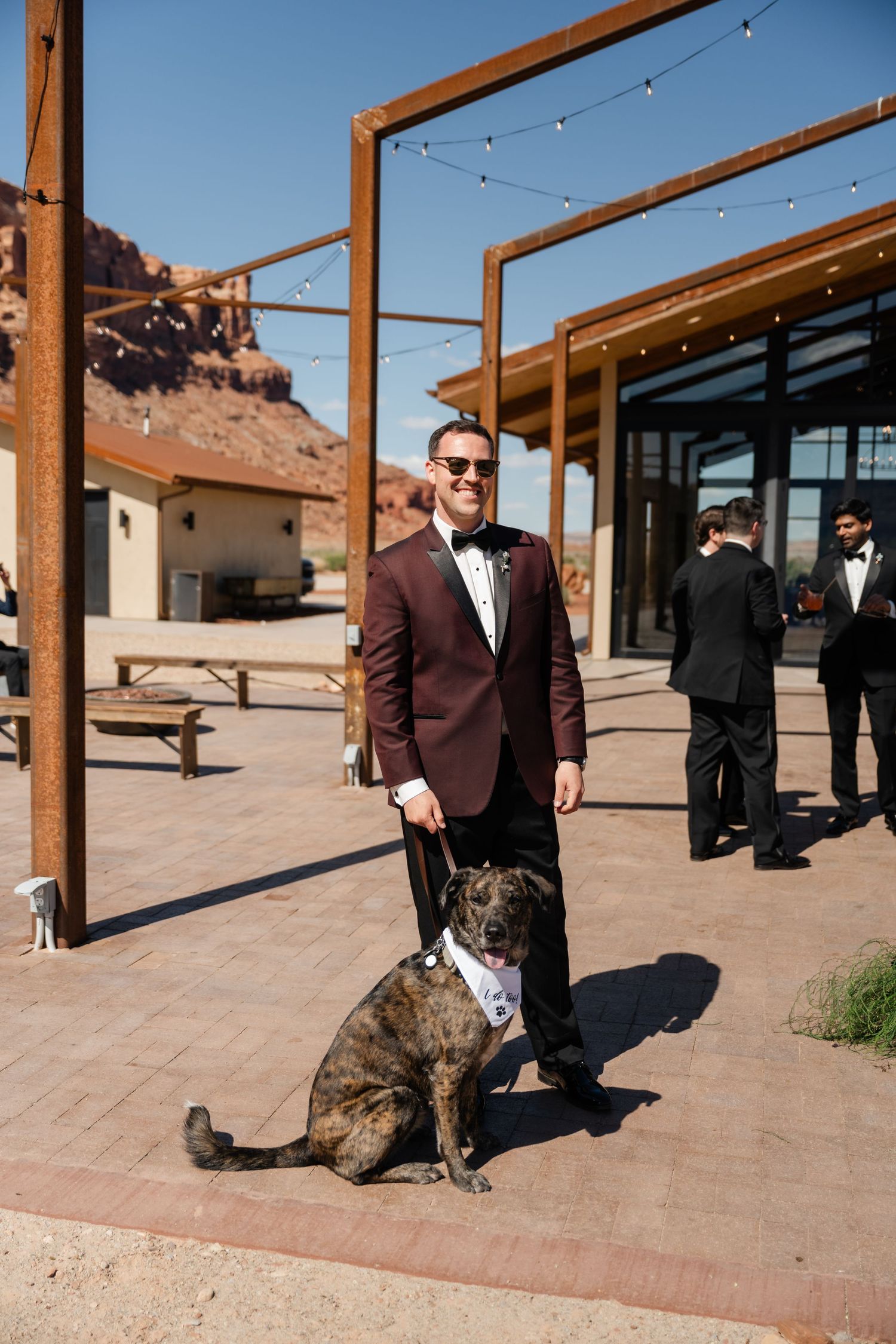 Man in burgundy tuxedo with brindle dog wearing bow tie at outdoor wedding venue with red rock backdrop.